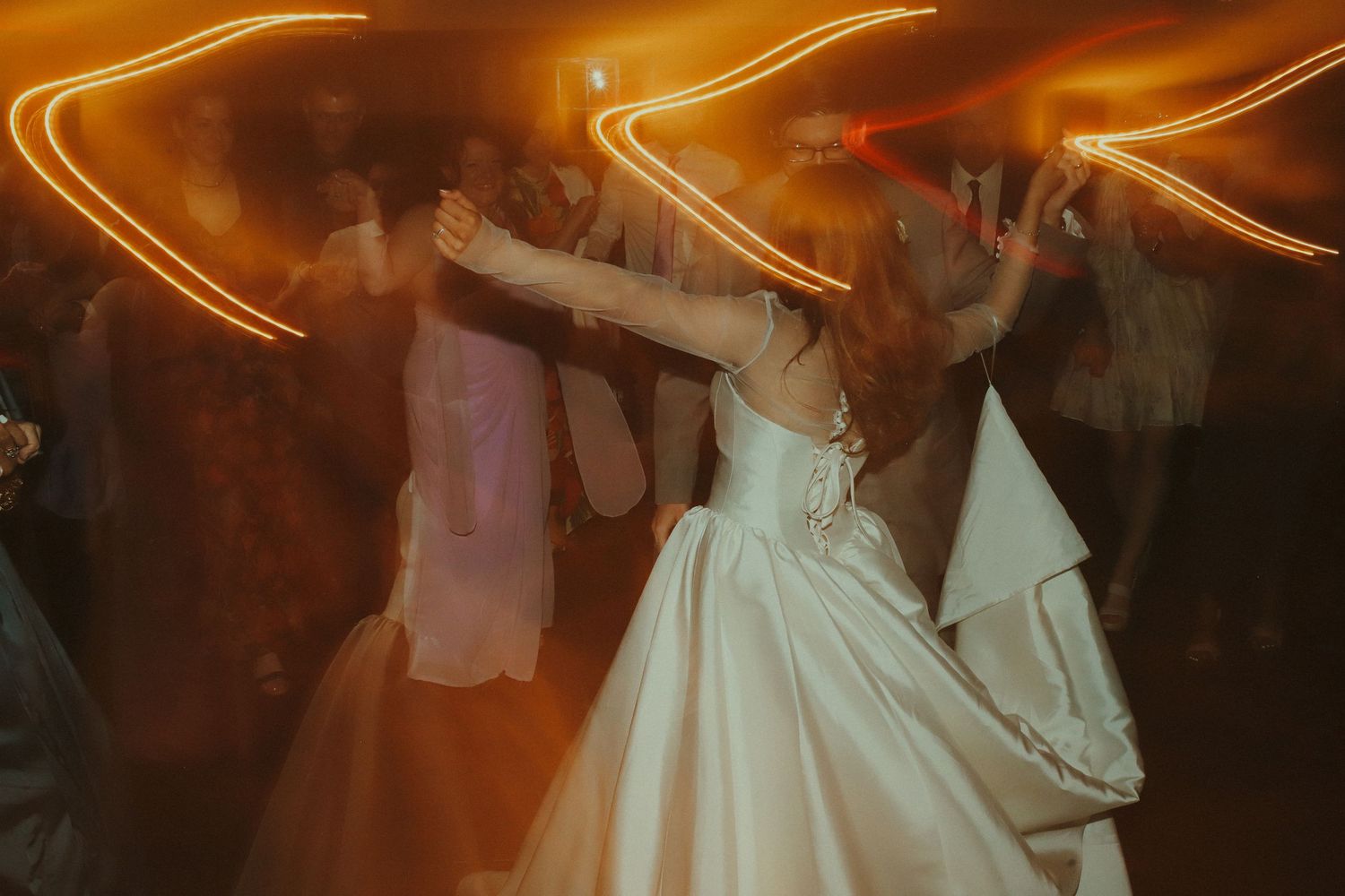 Bride dancing joyfully at wedding reception with motion blur light trails creating golden streaks in background.