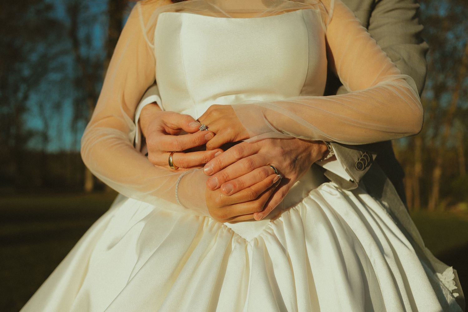 Bride and groom embracing, showing wedding rings, bride in white satin gown with sheer sleeves outdoors.