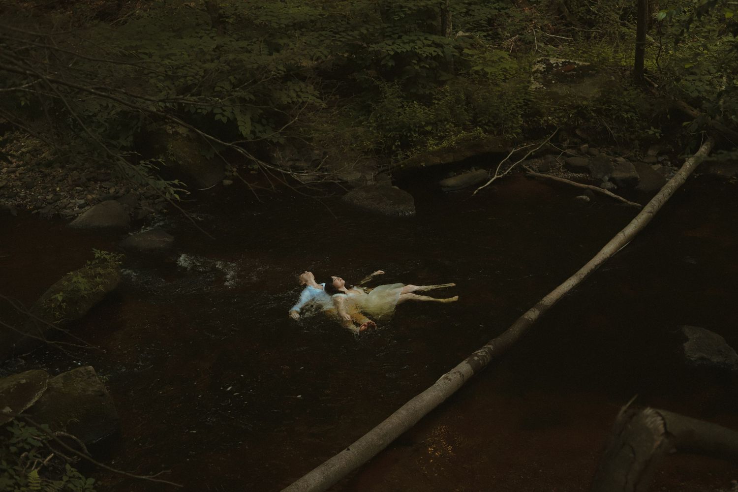 A person in a blue and white dress floating in a dark forest stream surrounded by rocks and fallen logs.