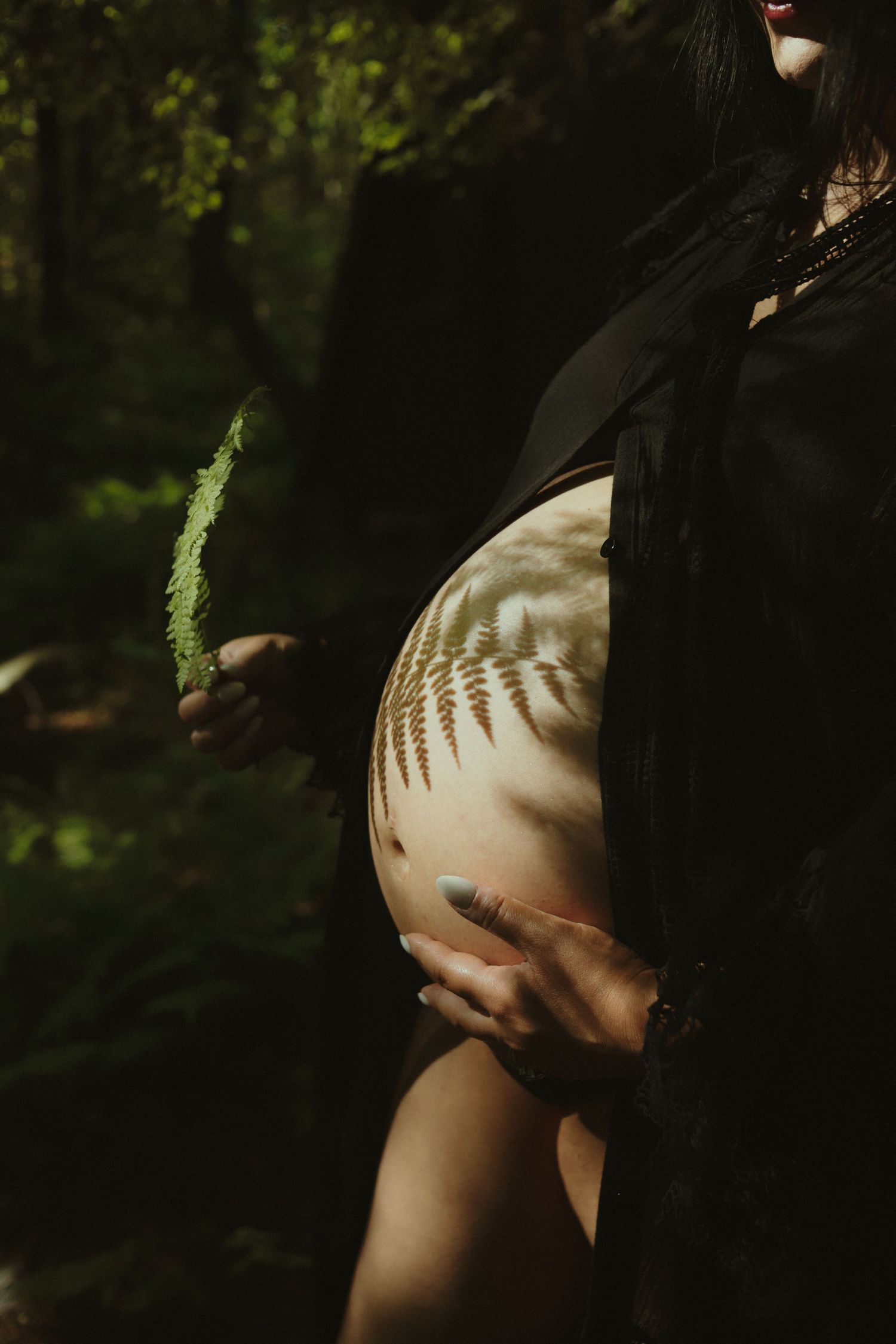 Pregnant woman in black robe holding fern leaf with fern shadow cast on bare belly in forest.