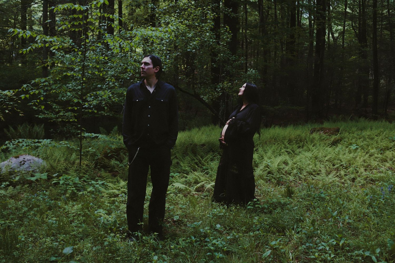 Two people standing in a lush green forest clearing, one in dark suit, one in black dress, surrounded by tall trees.