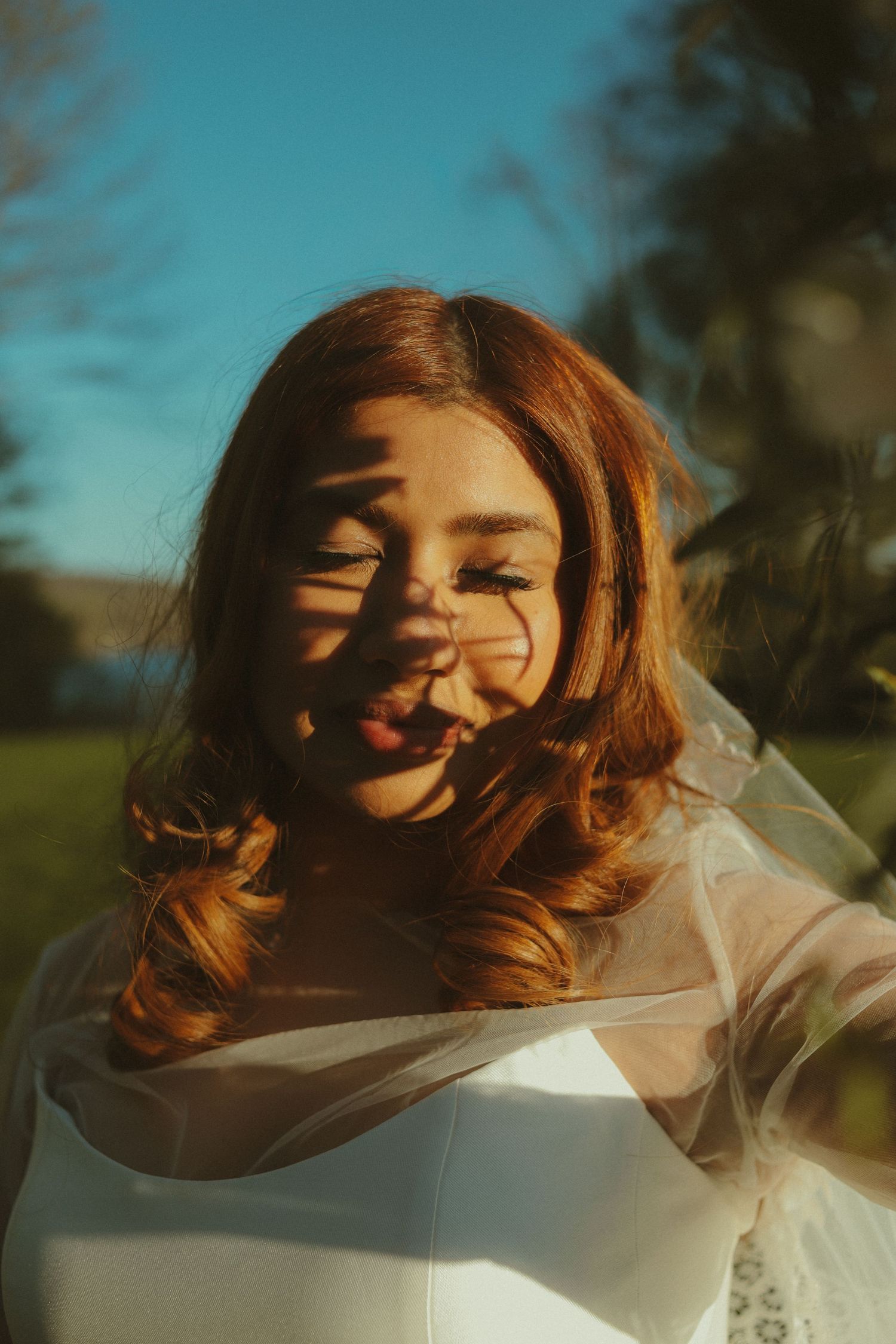 Bride in white dress with veil, auburn hair, playful expression, dappled sunlight shadows on face, blue sky background.