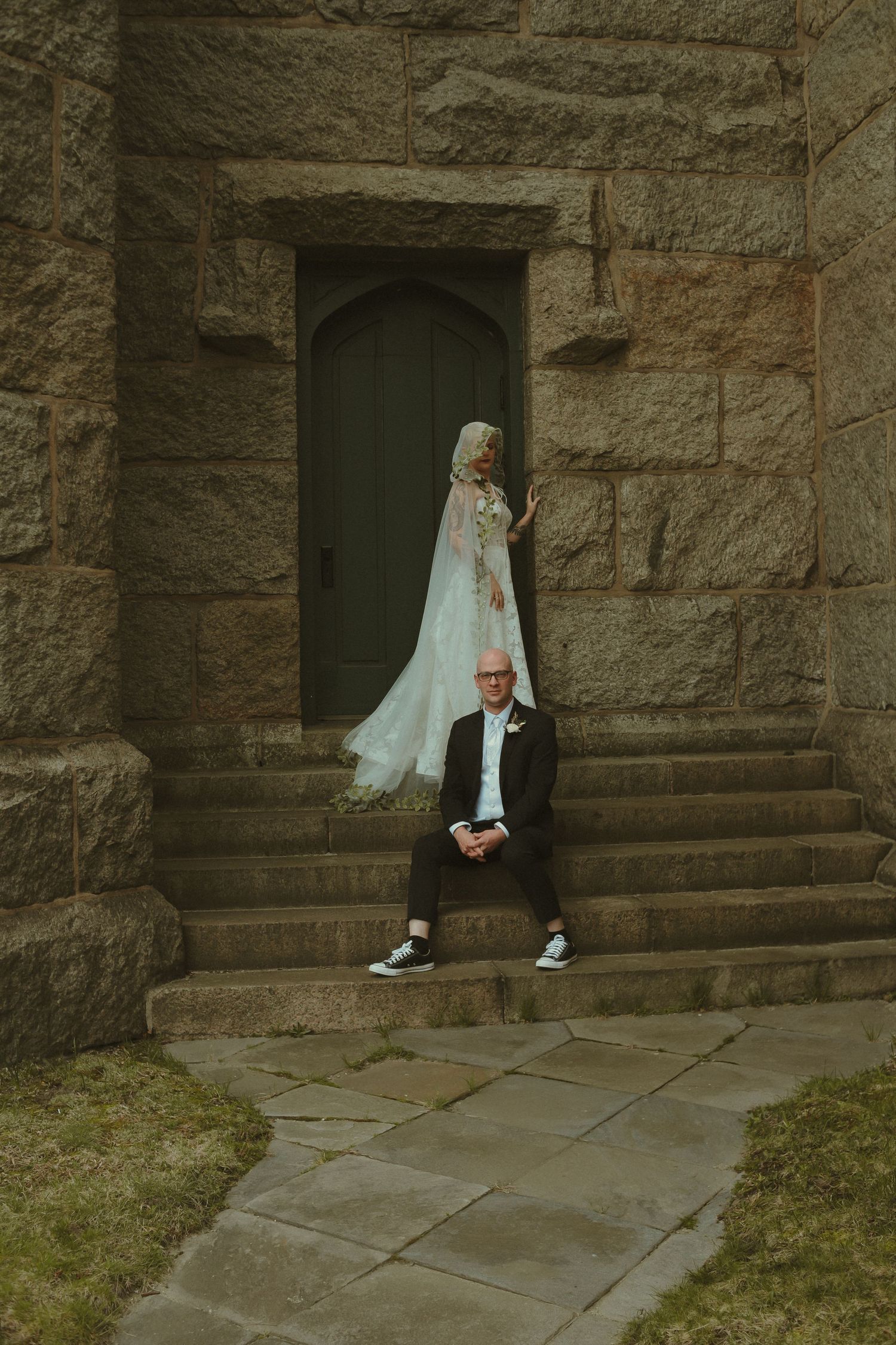 Bride in flowing veil and groom in dark suit posing on stone steps outside a rustic granite chapel door.