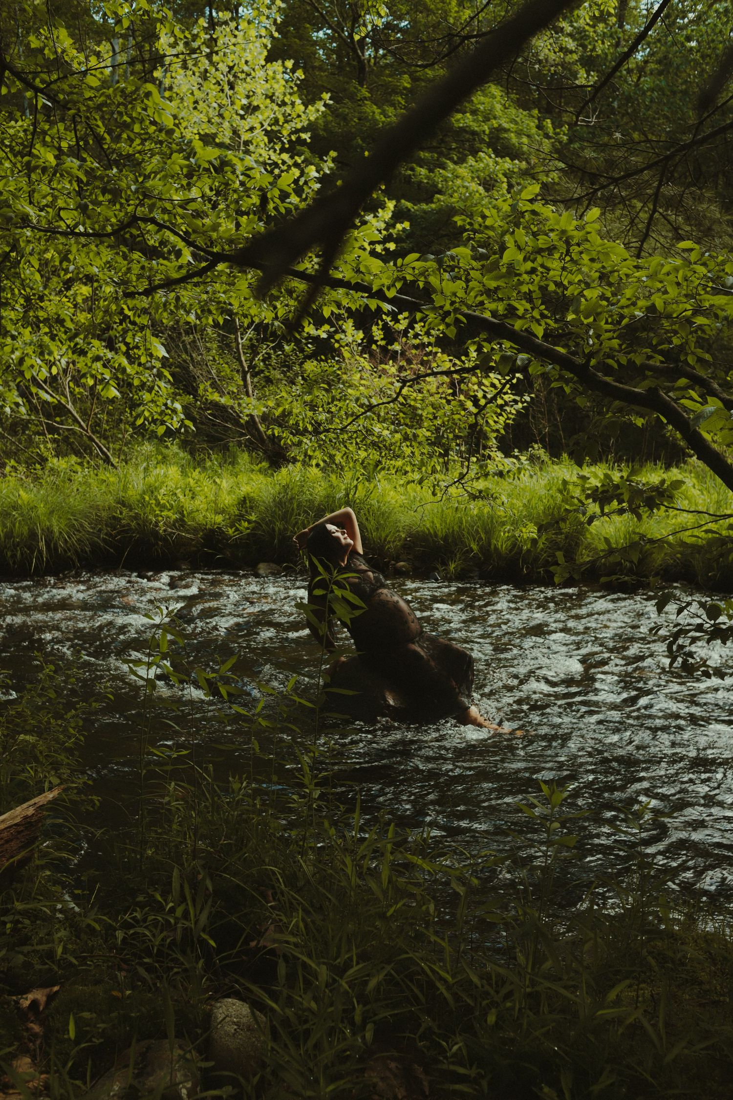 Person wading in a lush green forest stream surrounded by trees and flowing water.