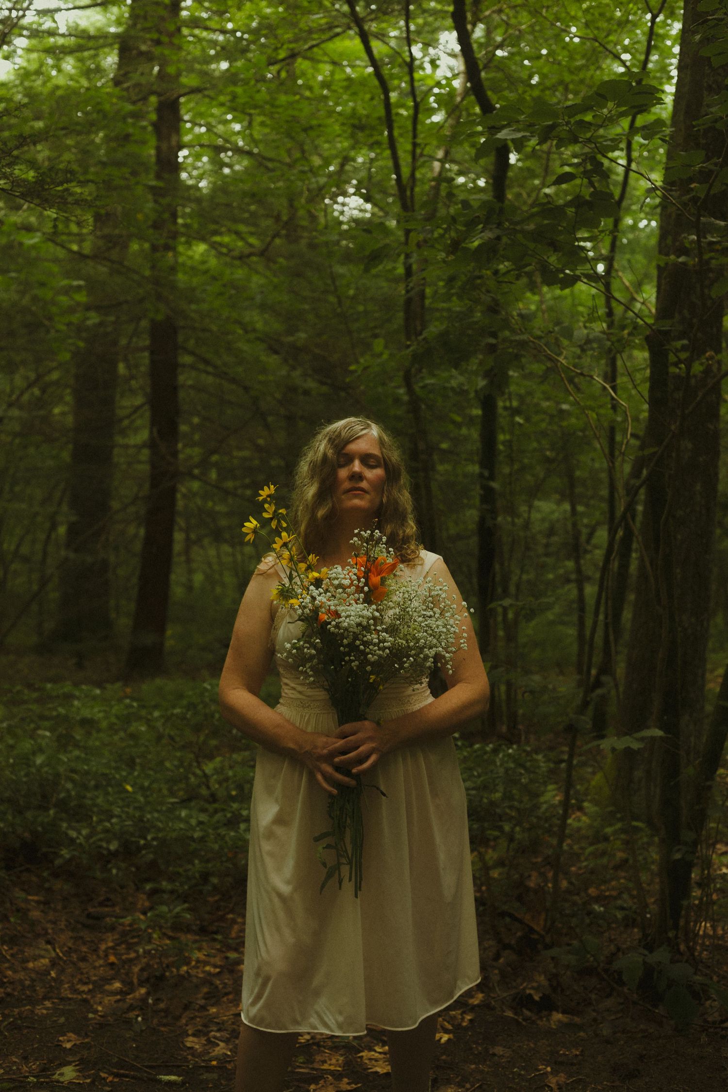 A person in a white dress holds a rustic wildflower bouquet while standing in a lush green forest setting.