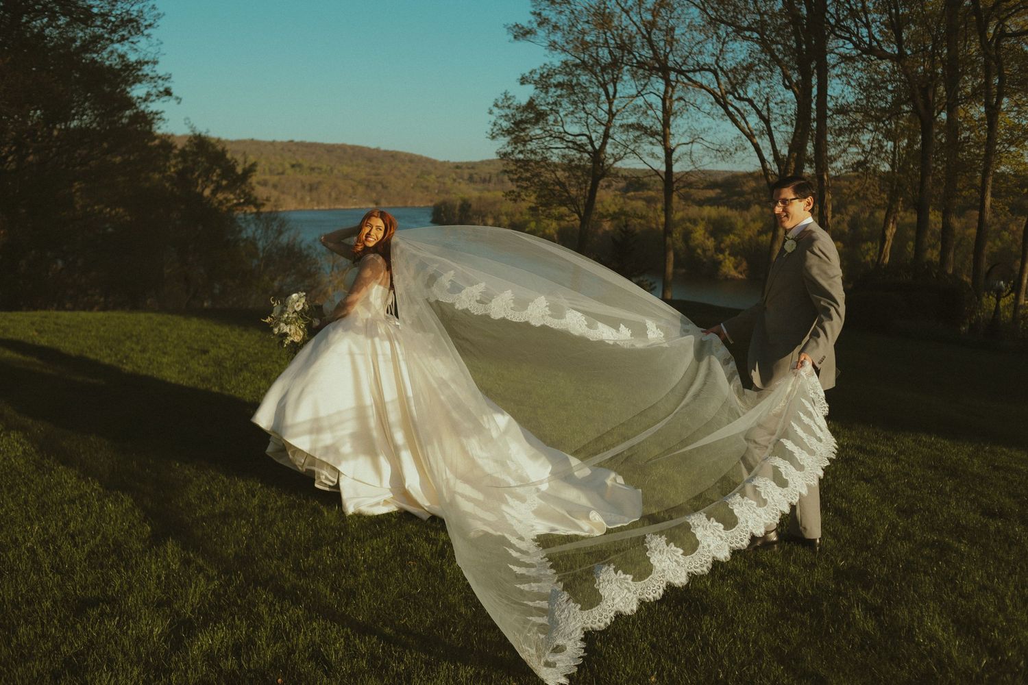 Bride and groom pose outdoors with flowing lace wedding veil billowing in the wind near a scenic lake.