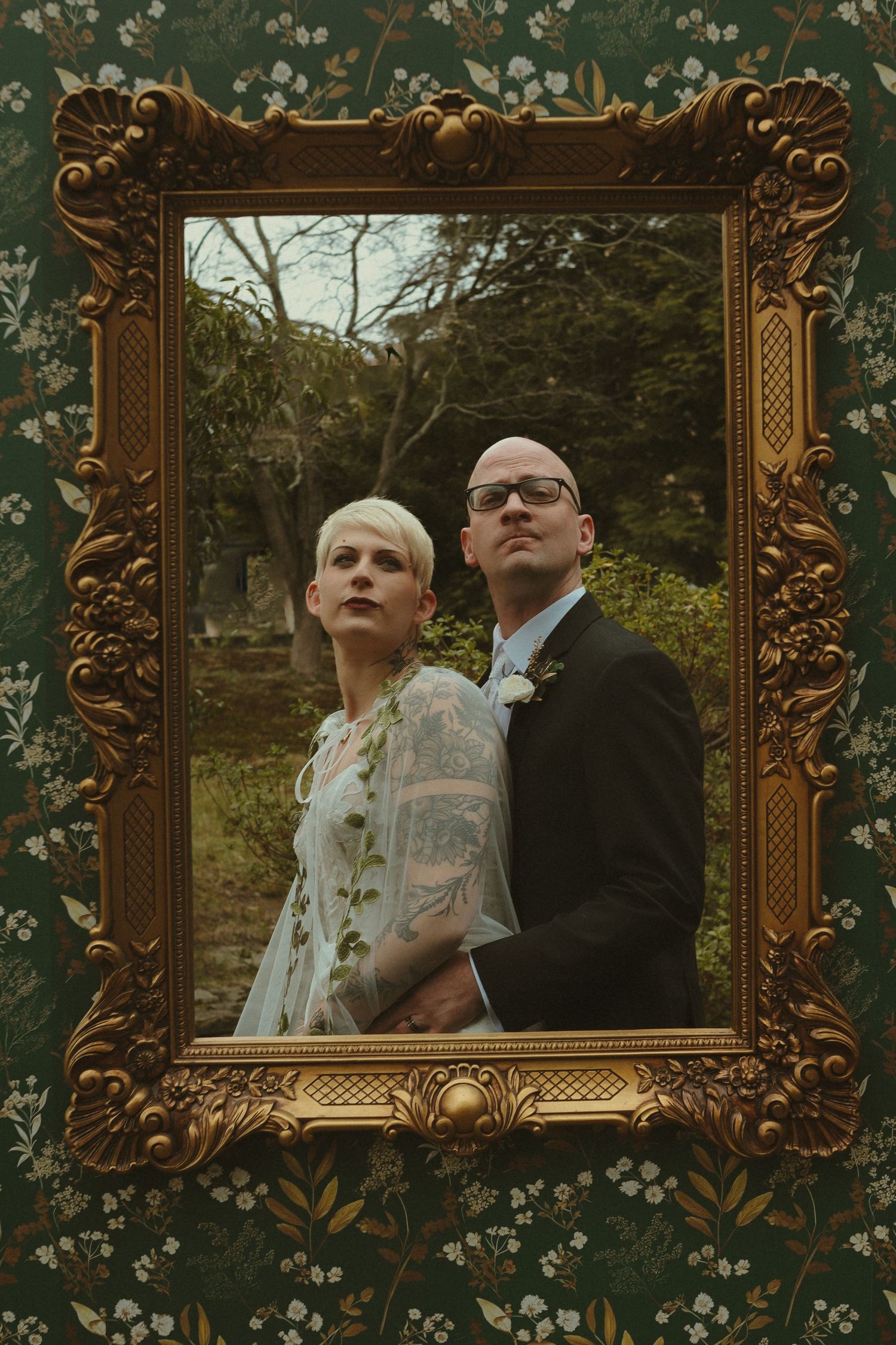 Bride and groom posing inside ornate gold picture frame against floral wallpaper in artistic wedding portrait.