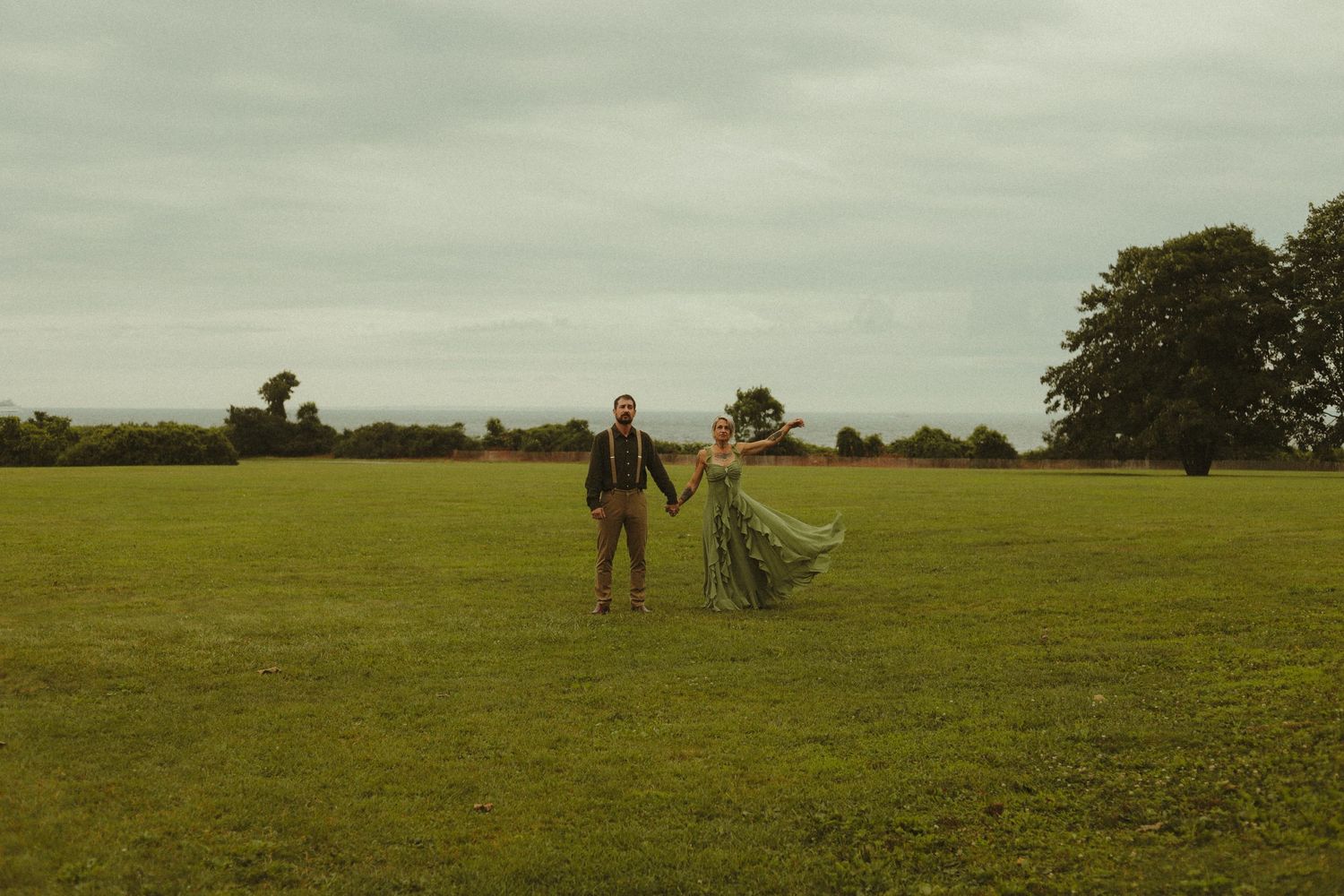 Couple holding hands in a vast green field, woman in flowing green dress, overcast sky, trees in background.