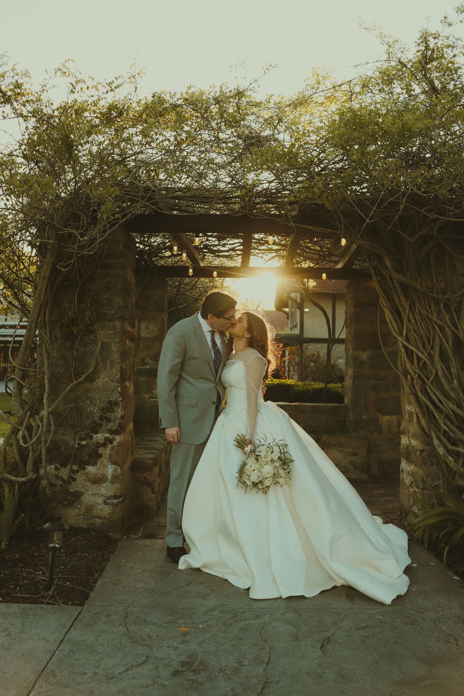 Bride and groom share a kiss under a rustic stone archway with vines at golden hour sunset.