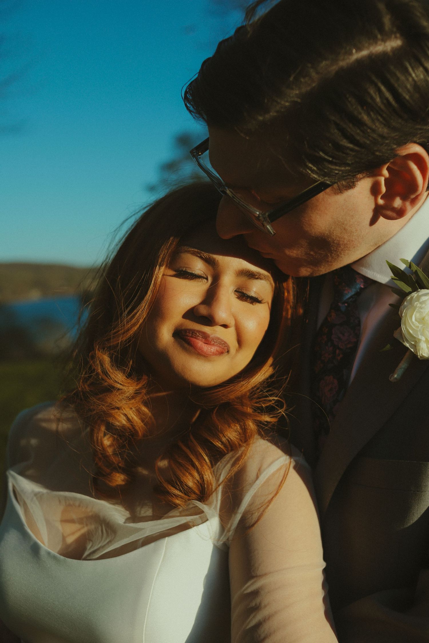 Groom in suit kissing bride on cheek outdoors, bride smiling with eyes closed in golden sunlight.