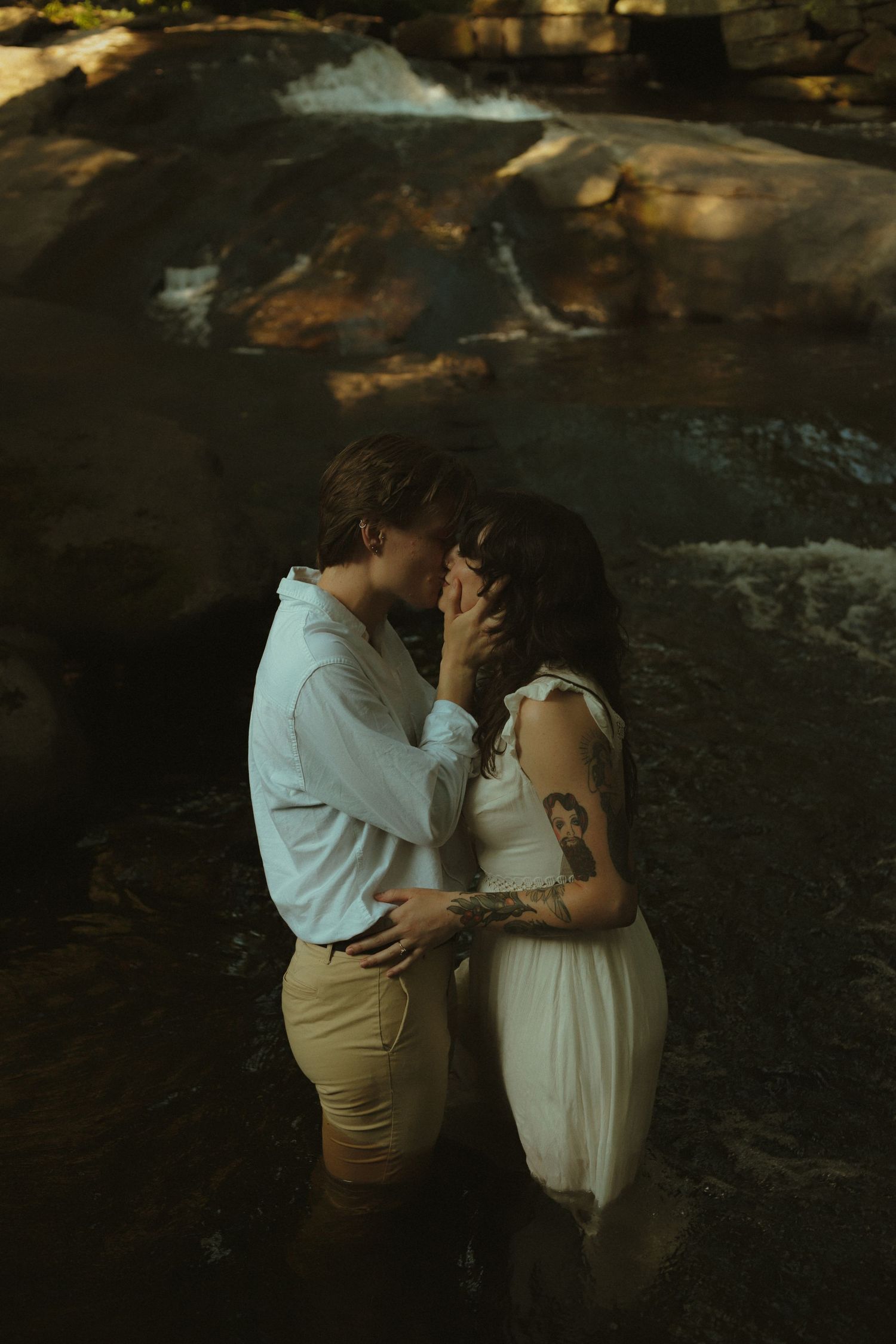 Couple sharing a romantic almost-kiss while standing in a rocky stream, woman in white dress with tattoos, man in white shirt.