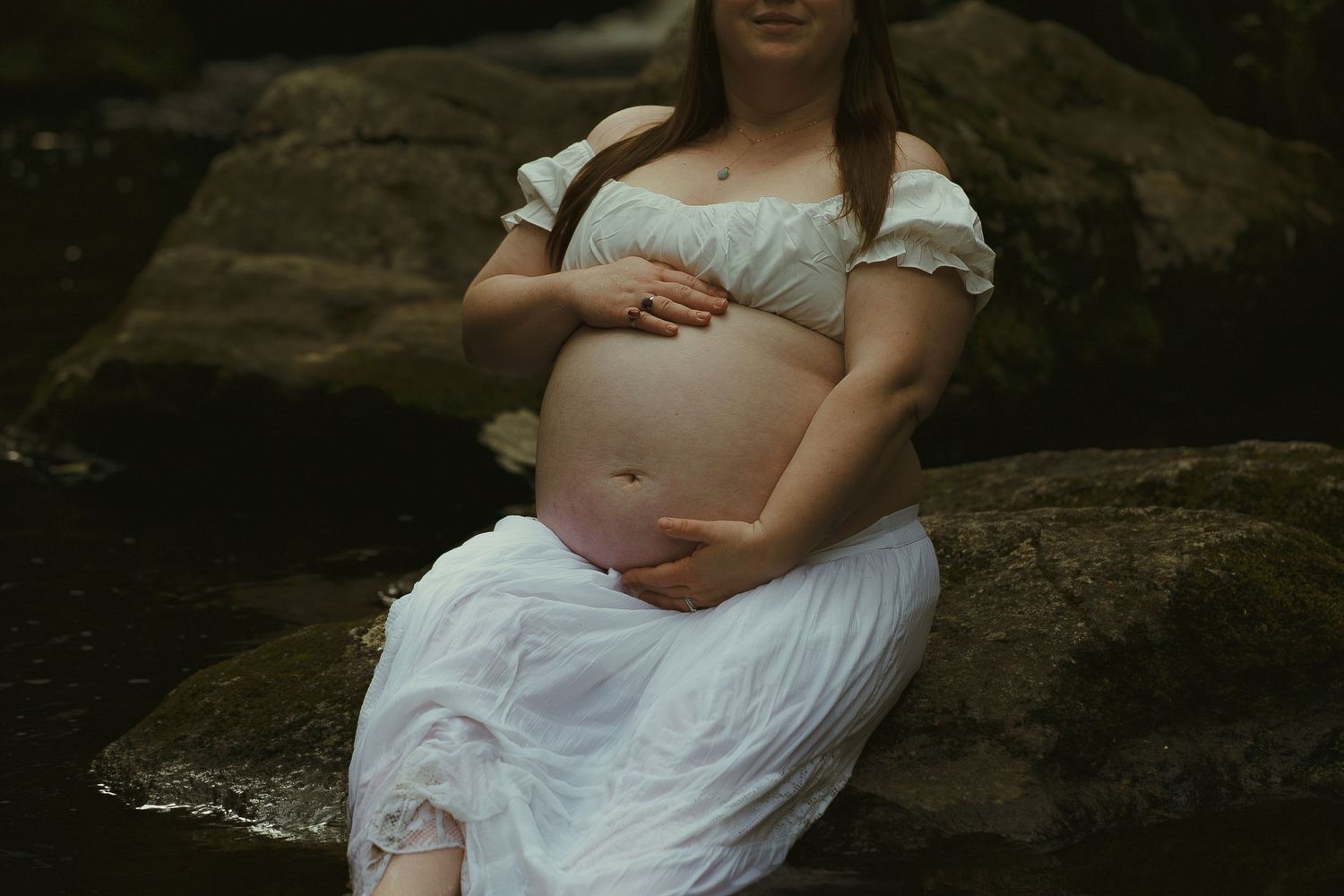 Pregnant woman in white dress sitting on rocks near water, cradling her baby bump during maternity photo session.