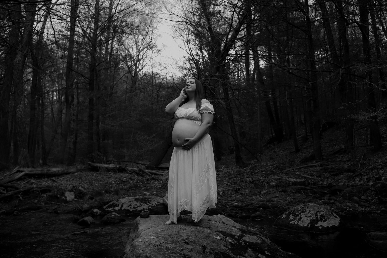 Black and white maternity photo of pregnant woman in white gown standing on rocks in a dark forest.