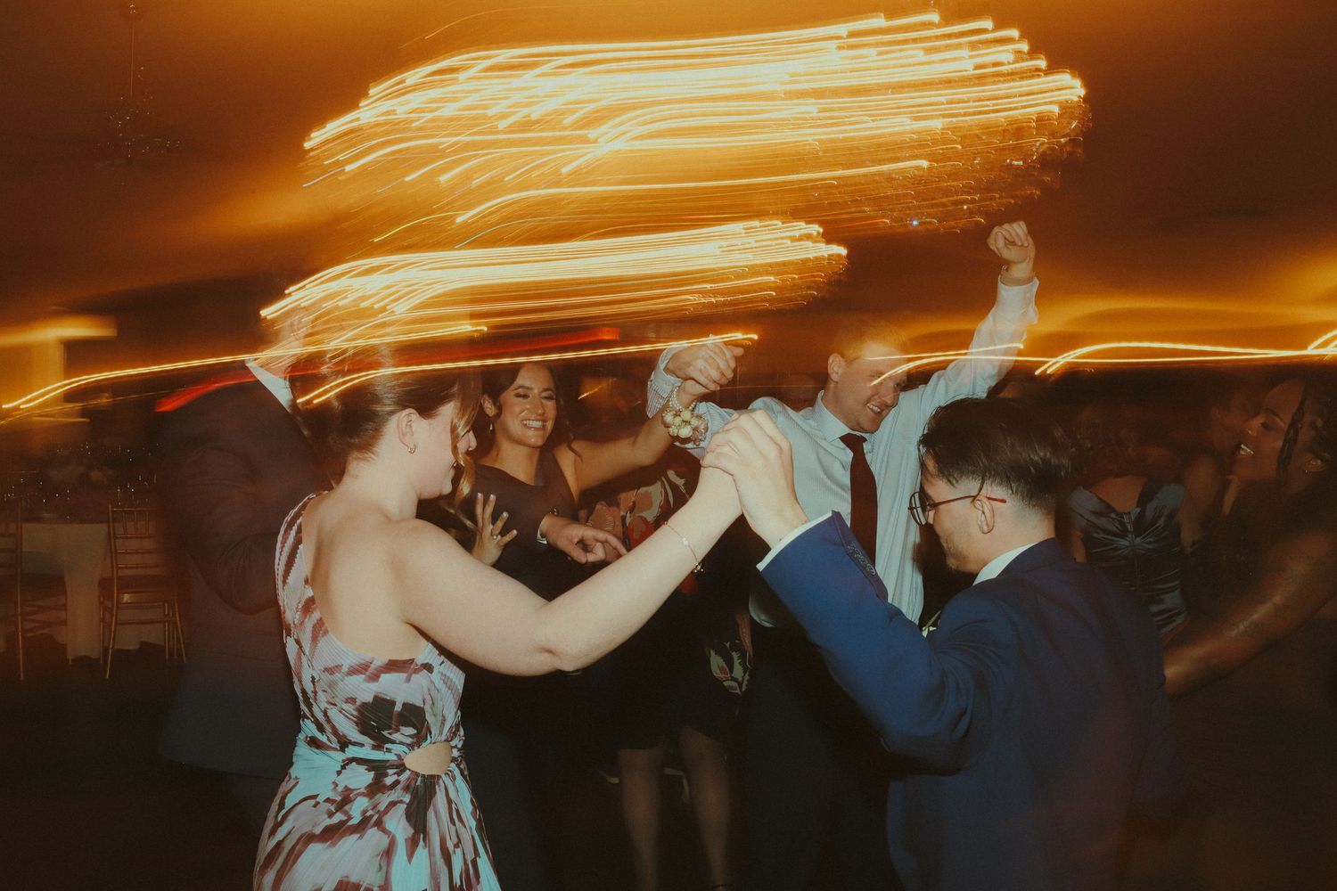Guests dancing joyfully at a wedding reception with warm light trails creating a festive atmosphere.