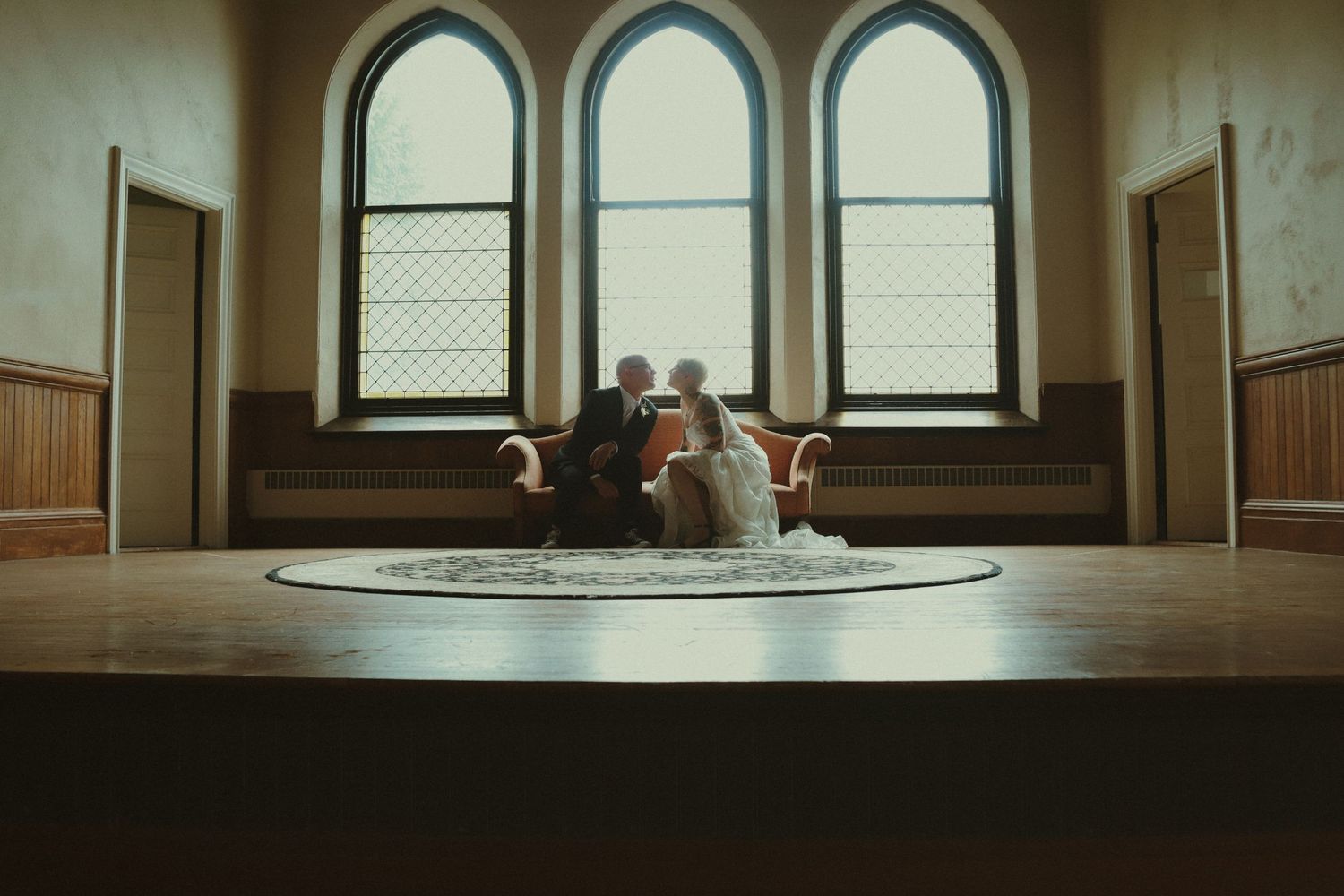 Newlyweds sitting intimately on vintage sofa in grand hall with arched windows during wedding day.