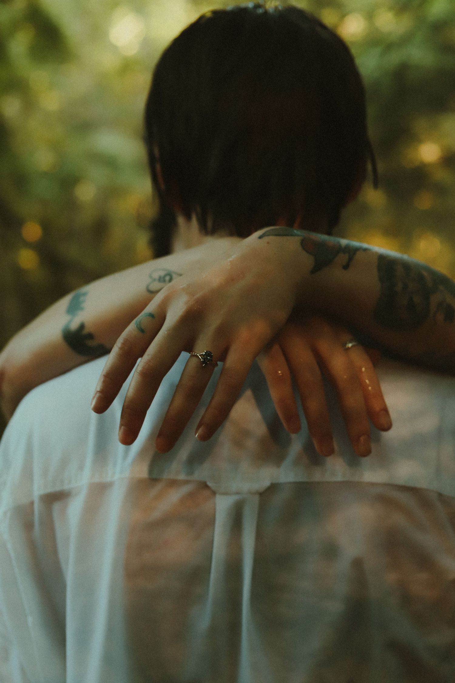 Tattooed arms of a couple embracing outdoors, with an engagement ring visible on one hand.