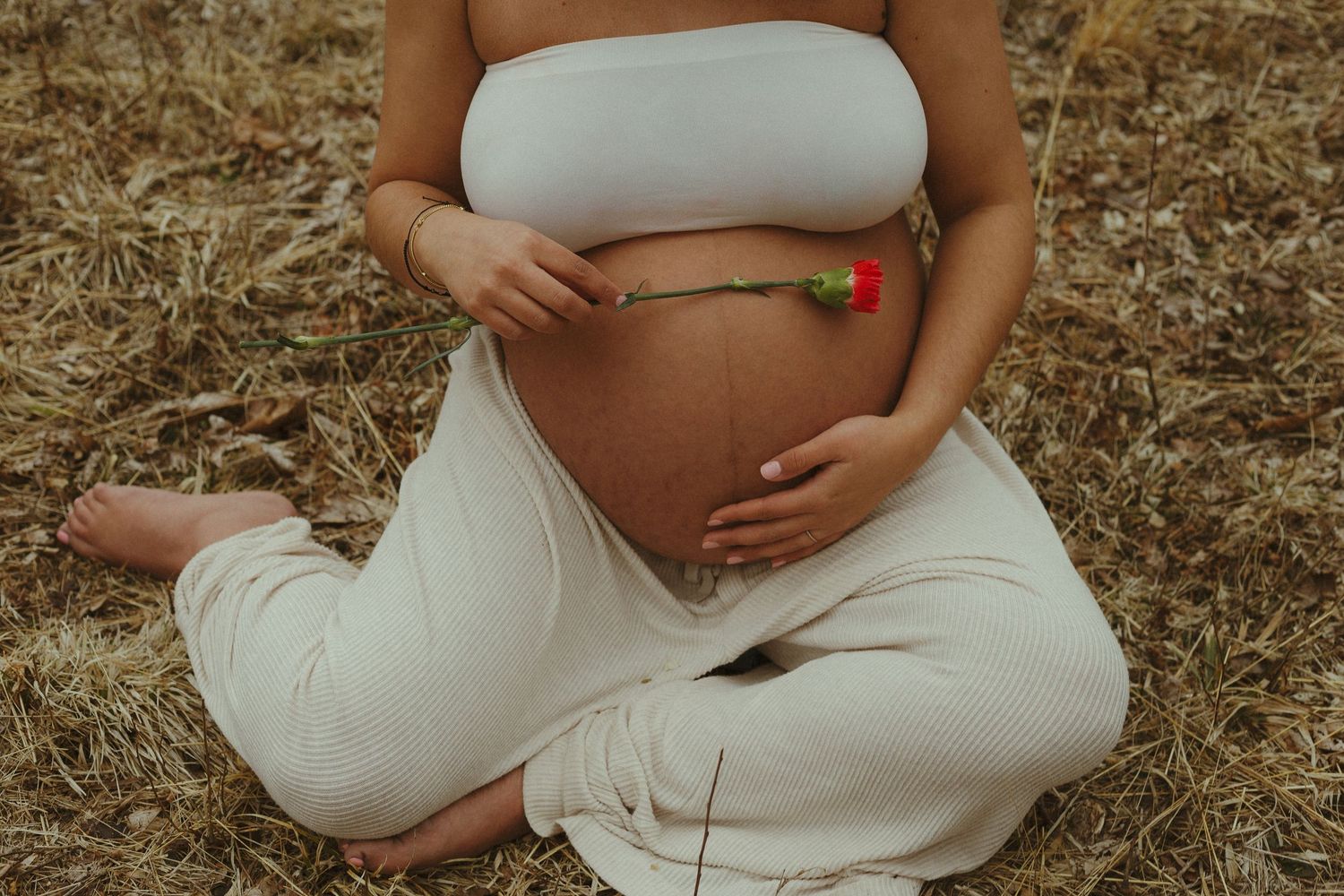 A person in white clothing sits cross-legged outdoors in a natural setting during a maternity photoshoot.
