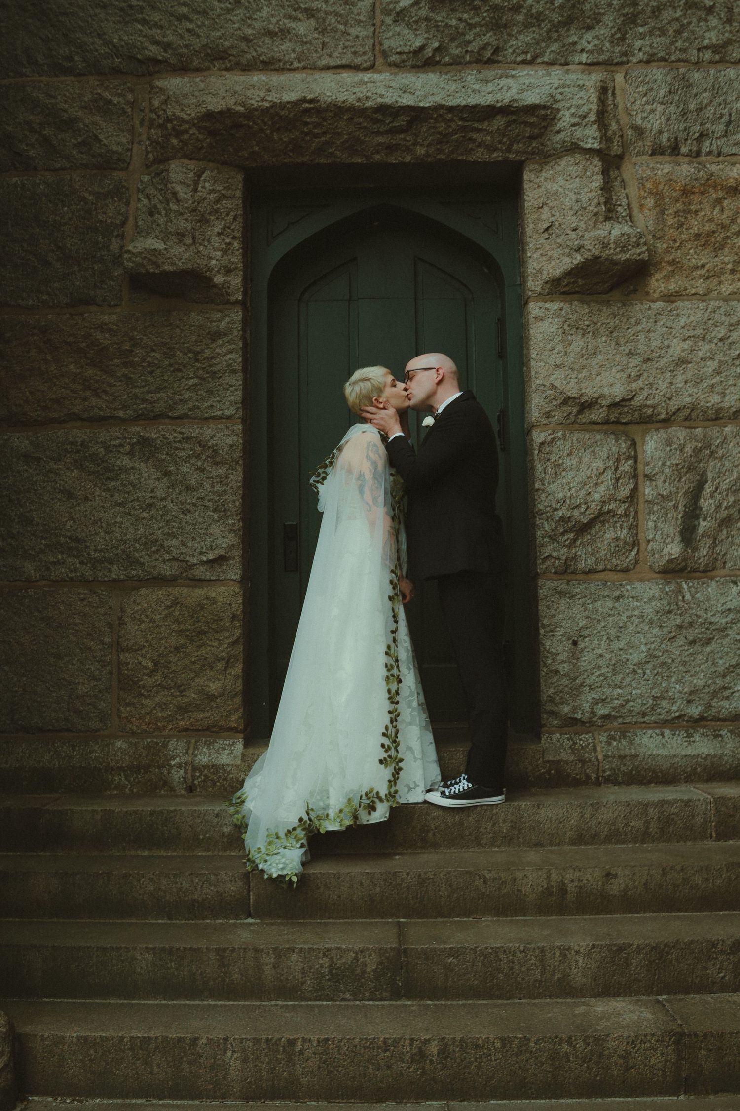 Bride and groom sharing a romantic kiss on stone church steps, bride wearing a flowing white gown with floral veil.