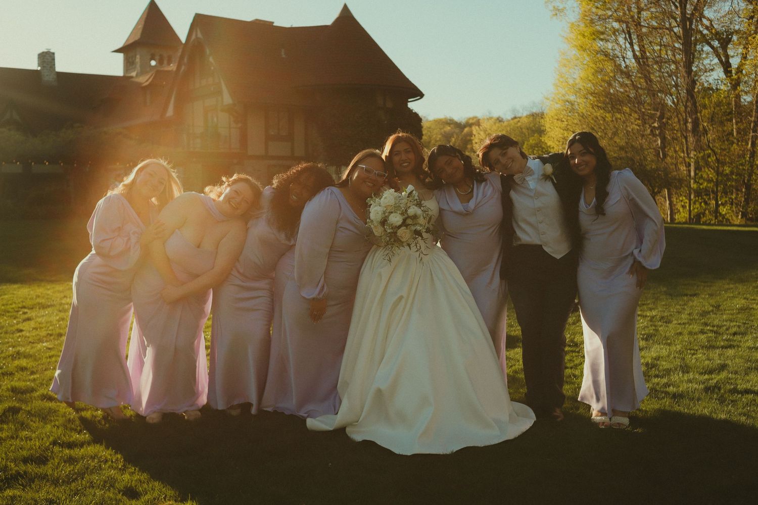 Bride in white gown poses with bridesmaids in pink dresses at golden hour sunset outside elegant estate.