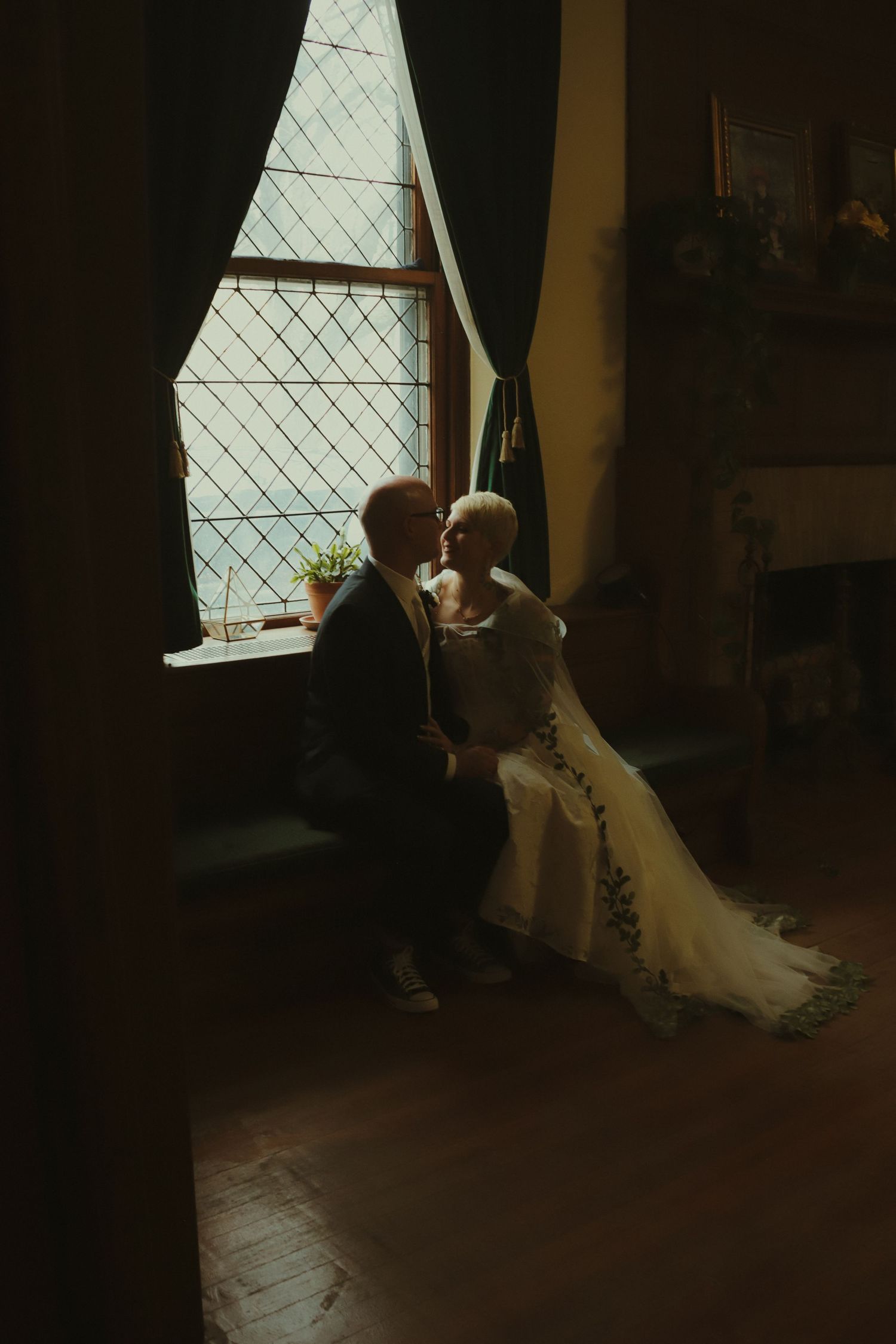 Bride and groom share a romantic moment seated by a leaded glass window in a dark room.