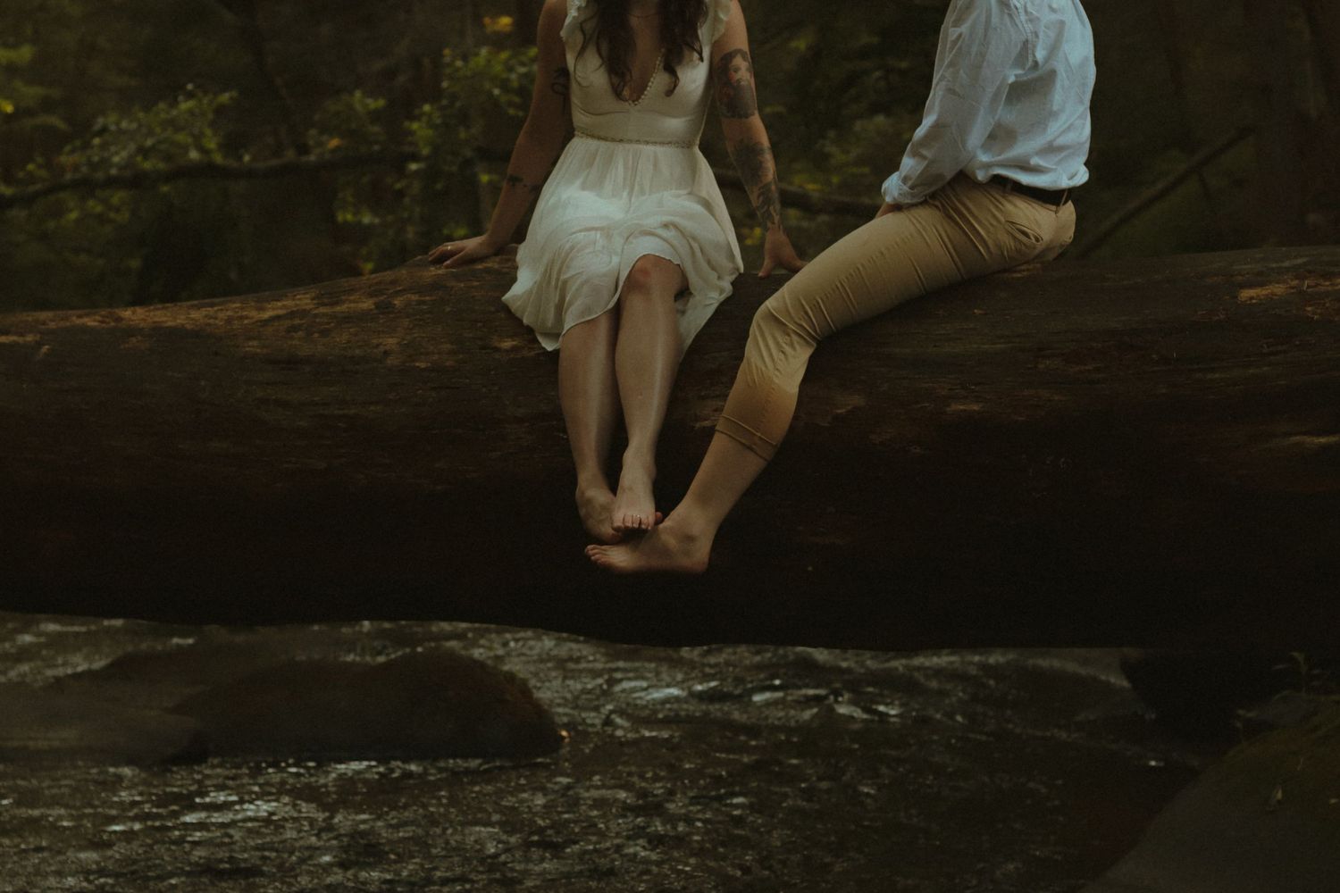 Couple sitting barefoot on a fallen log over a rocky stream in a lush forest setting.