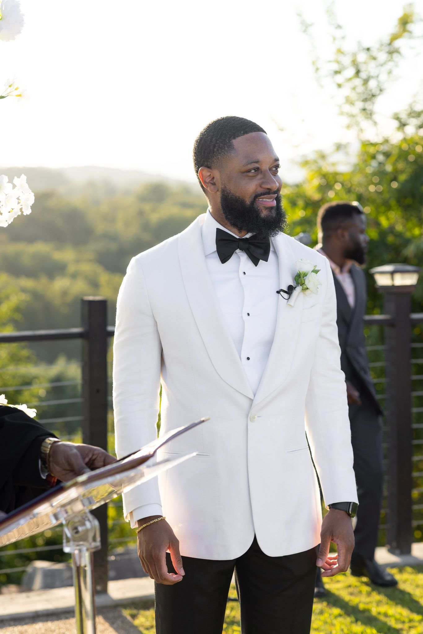 A groom in a white dinner jacket and black bow tie smiles during an outdoor wedding ceremony.