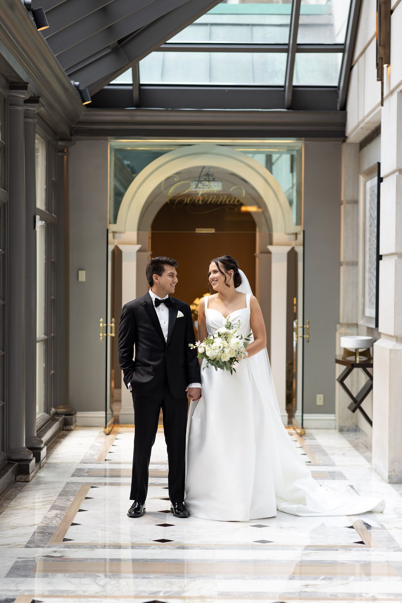 A wedding couple poses in an elegant hallway with marble floors and arched windows creating a romantic architectural setting.