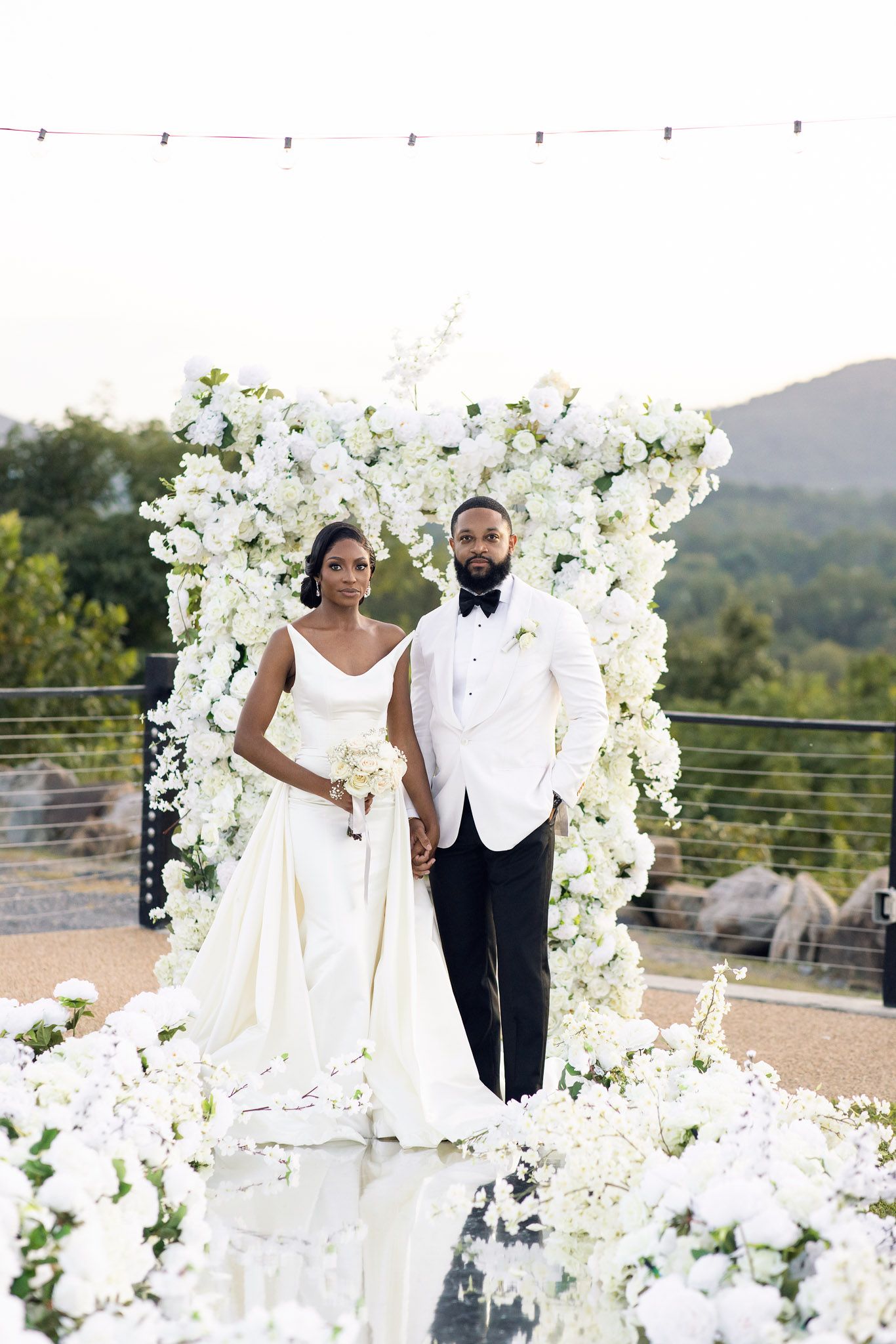 Elegant wedding portrait with couple standing on reflective floor under floral arch.