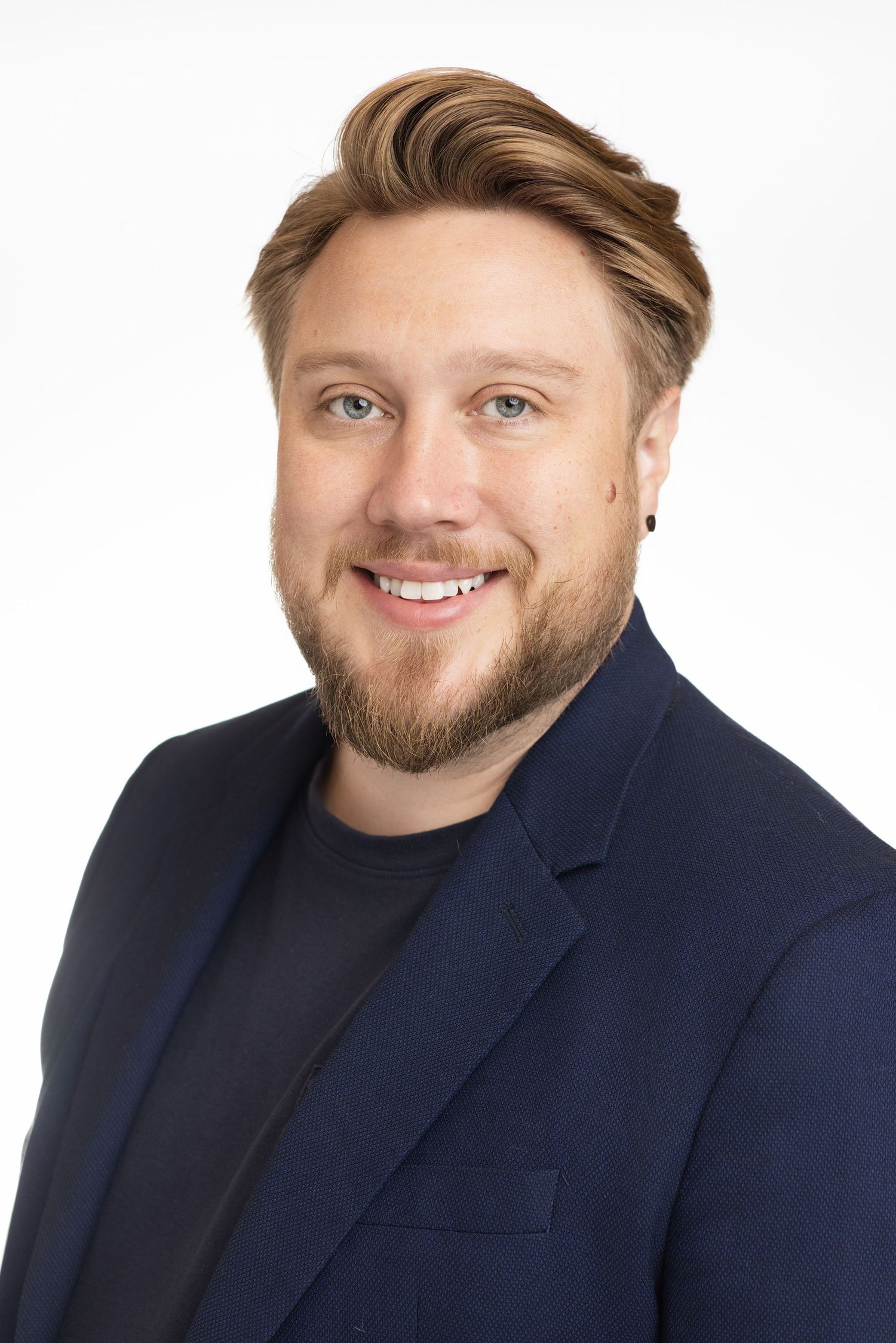 Smiling man with beard and blue eyes wearing a navy blazer poses for a professional headshot on white background.