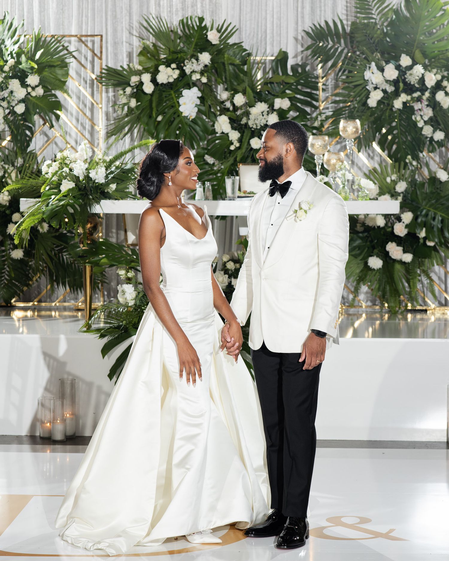 Elegant wedding couple in white dress and tuxedo pose together against a backdrop of lush greenery and white flowers.