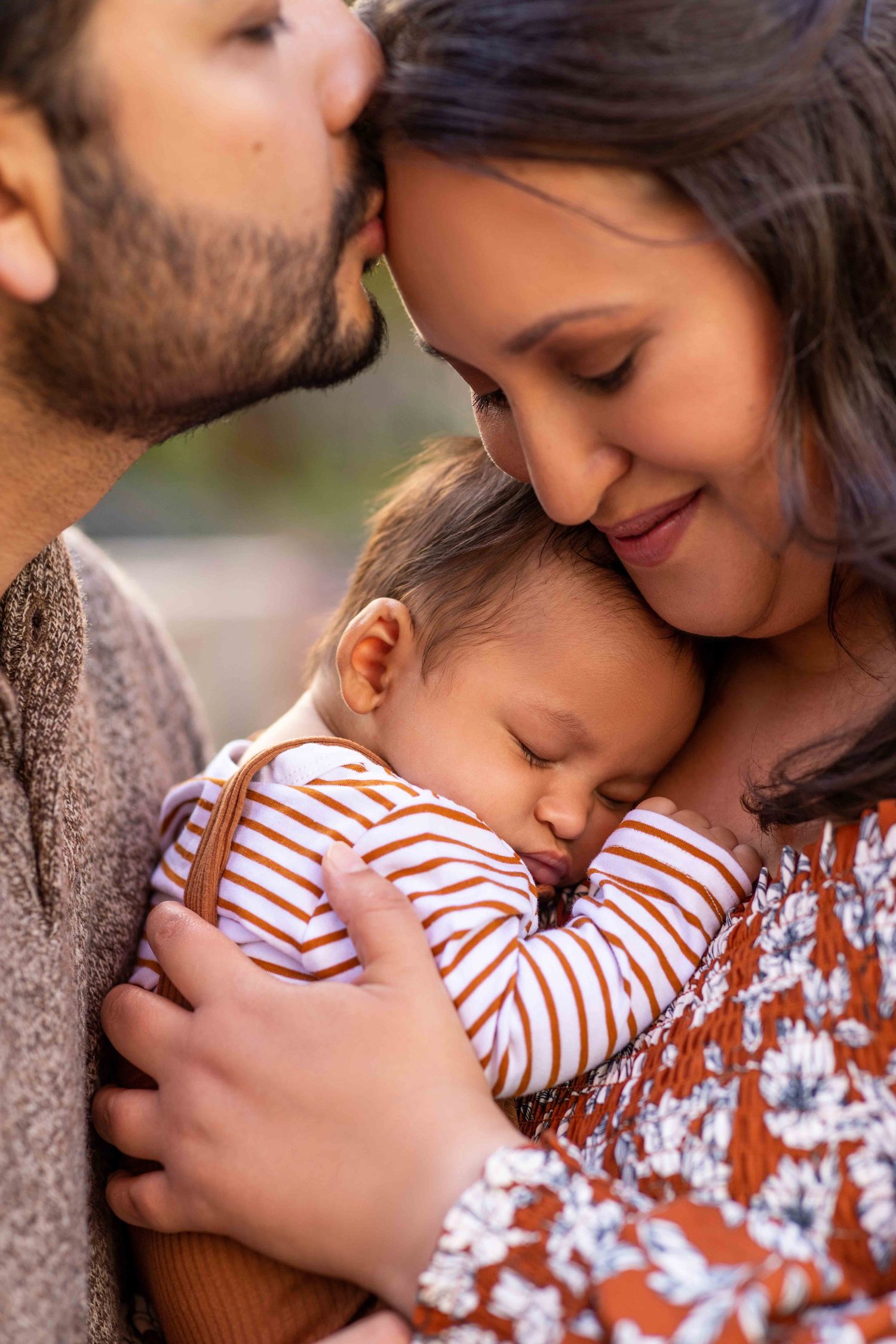 Parents tenderly embrace their sleeping newborn baby, sharing a loving family moment outdoors.