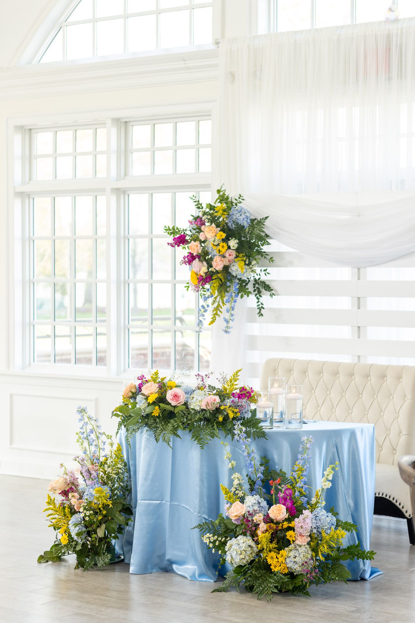 Elegant wedding reception tables with light blue tablecloths and colorful floral arrangements in a bright white venue.