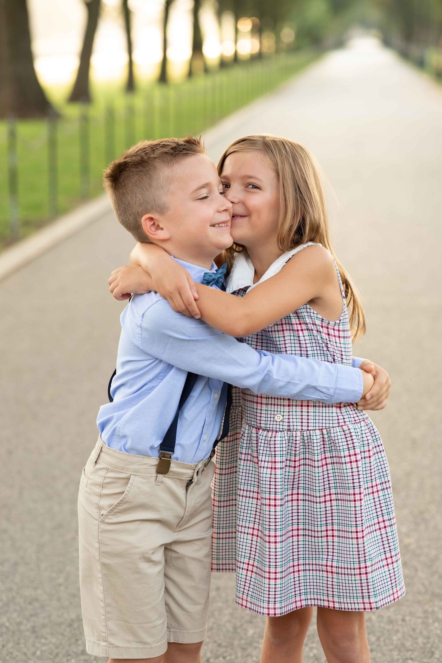 Two young children hugging and smiling on a tree-lined path at sunset, dressed in smart casual outfits.