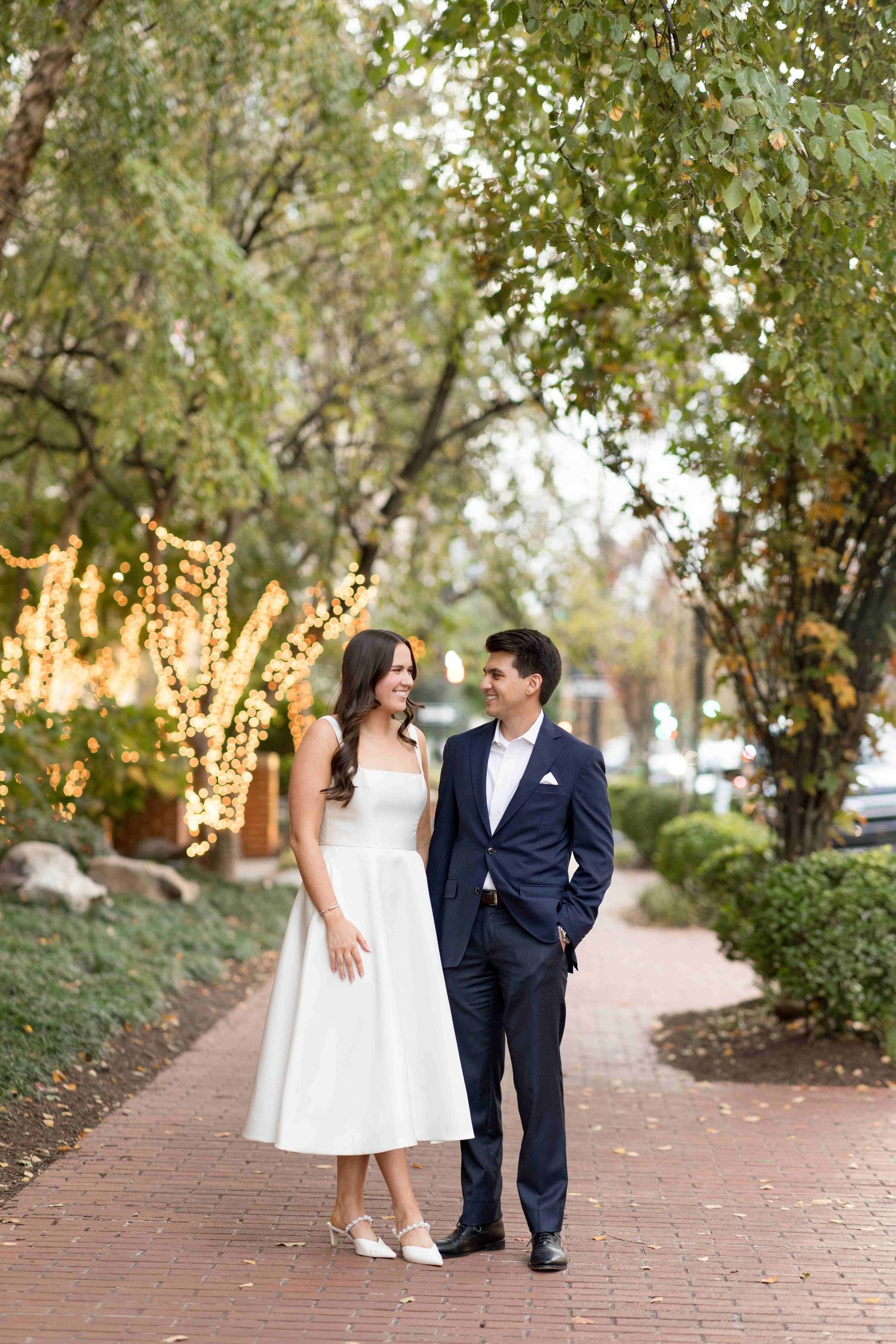 Couple in wedding attire walking on brick path surrounded by trees with fairy lights.
