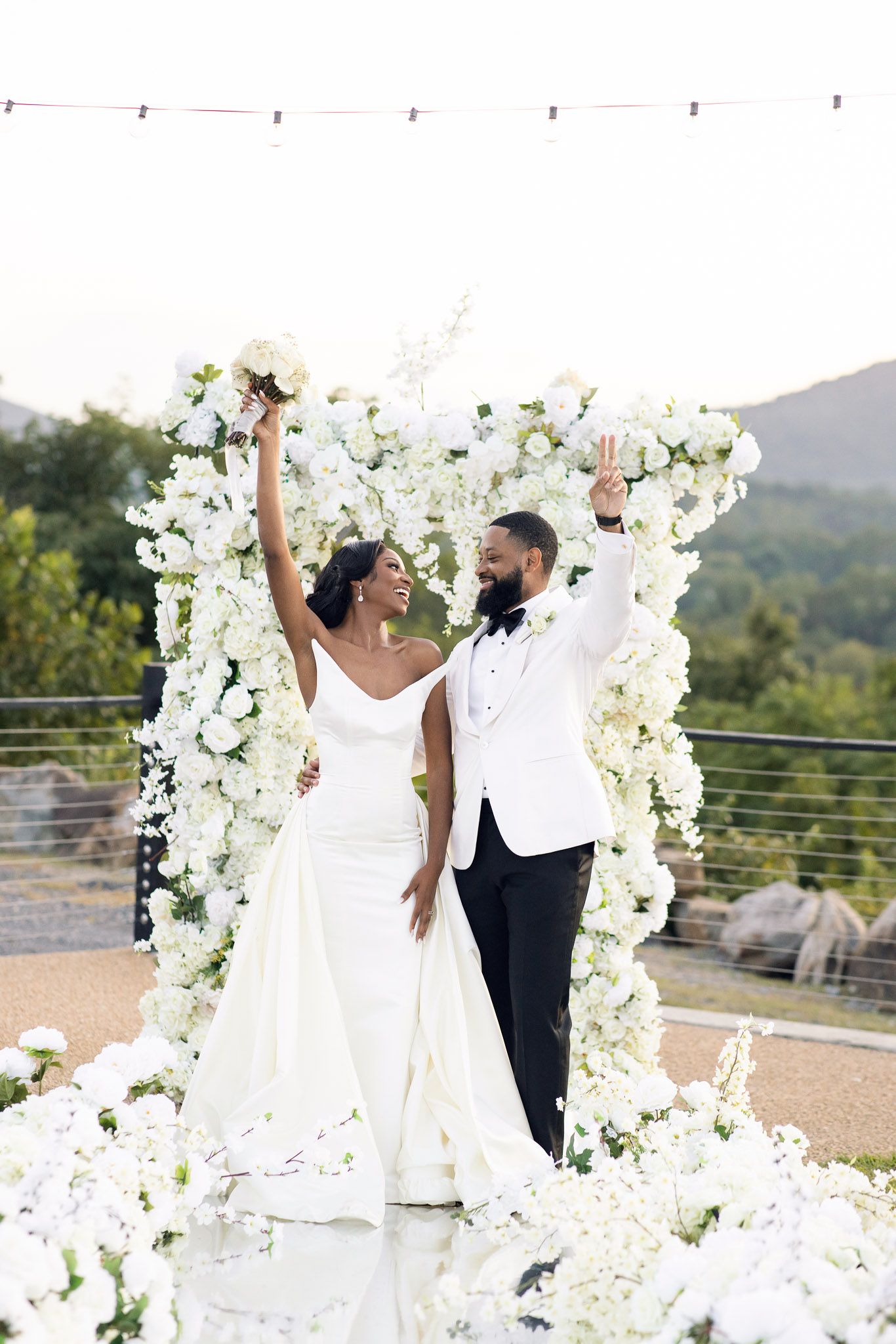 Celebratory wedding pose with couple raising arms under white flower arch.