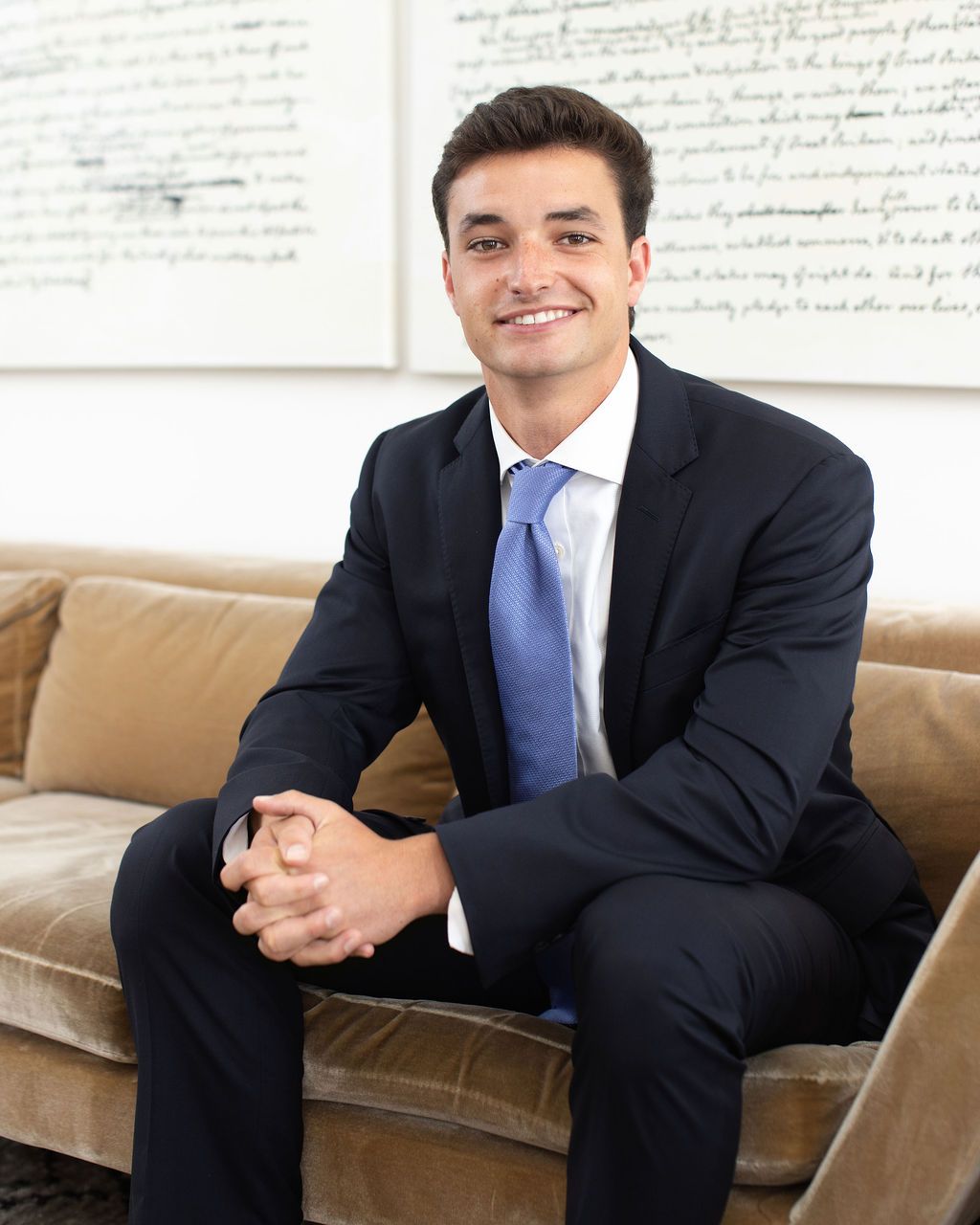 Young professional man in navy suit and blue tie sitting on beige velvet sofa in elegant office setting.