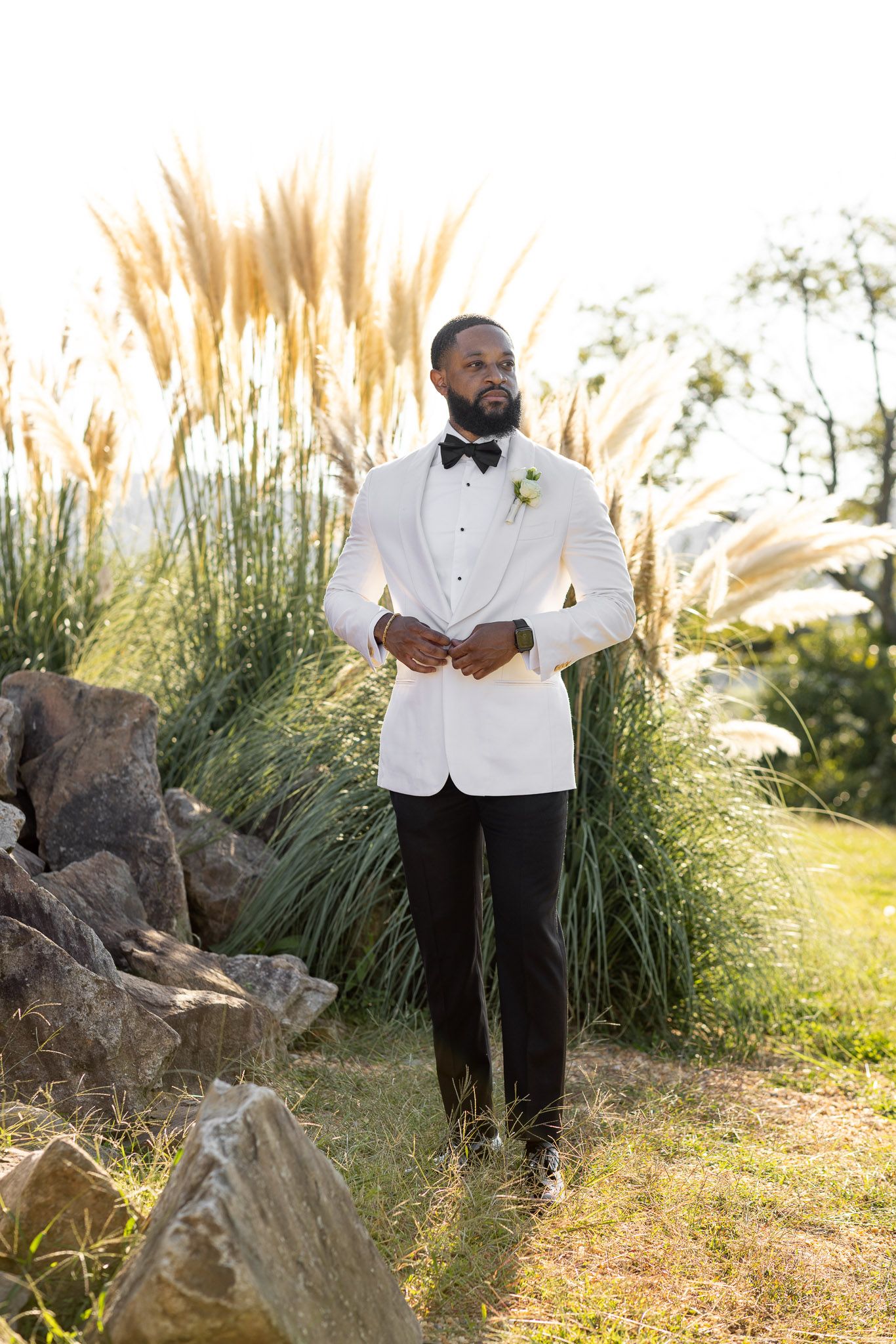 Elegant groom in white dinner jacket poses against ornamental grasses and rocks in outdoor setting.
