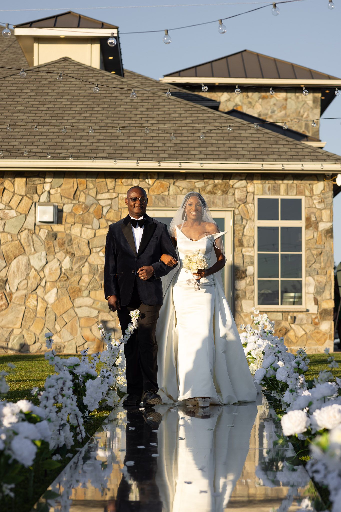 A wedding couple poses on a reflective aisle decorated with white flowers outside a stone building.