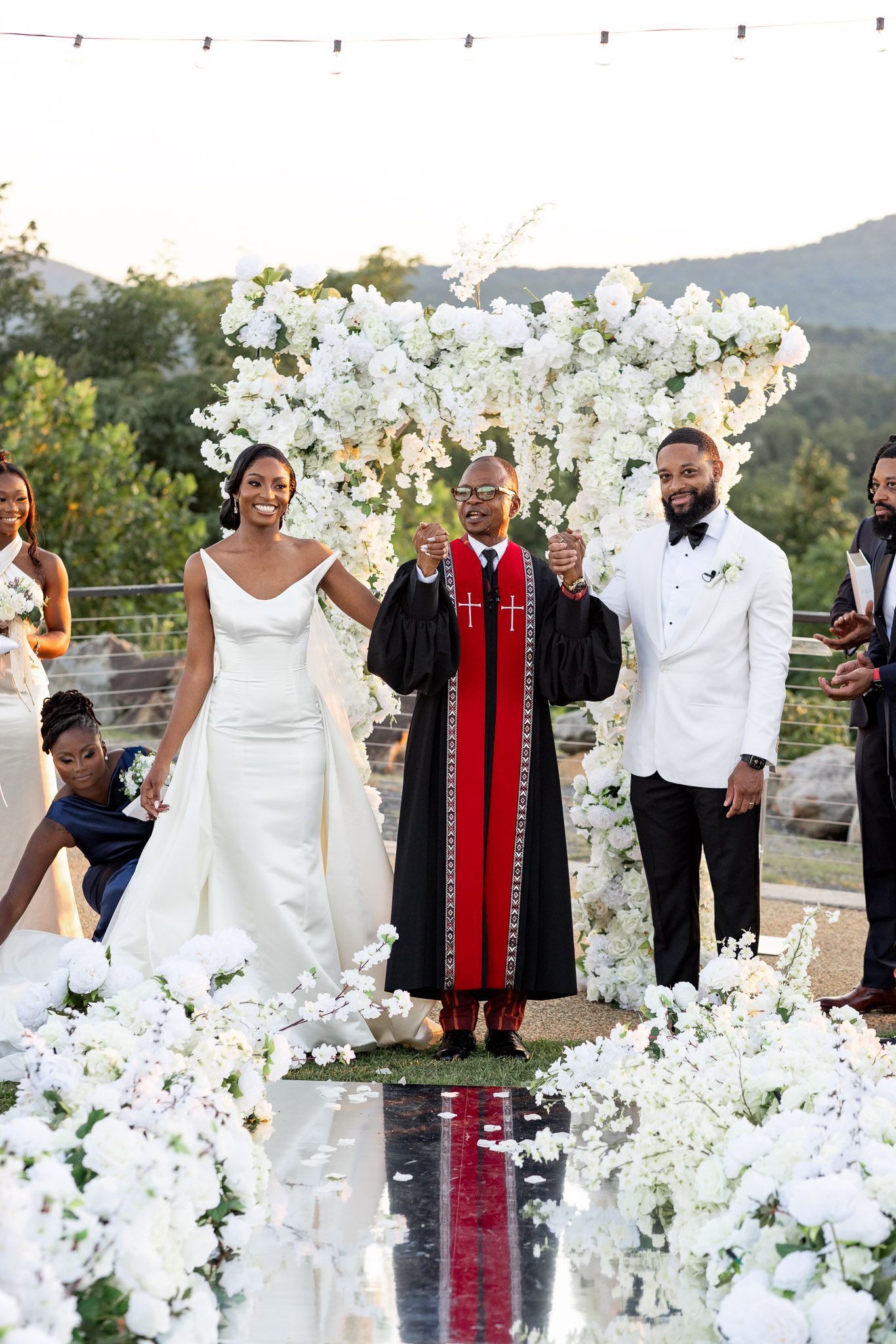 Wedding party gathered for ceremony beneath spectacular white floral arch with mountain views.