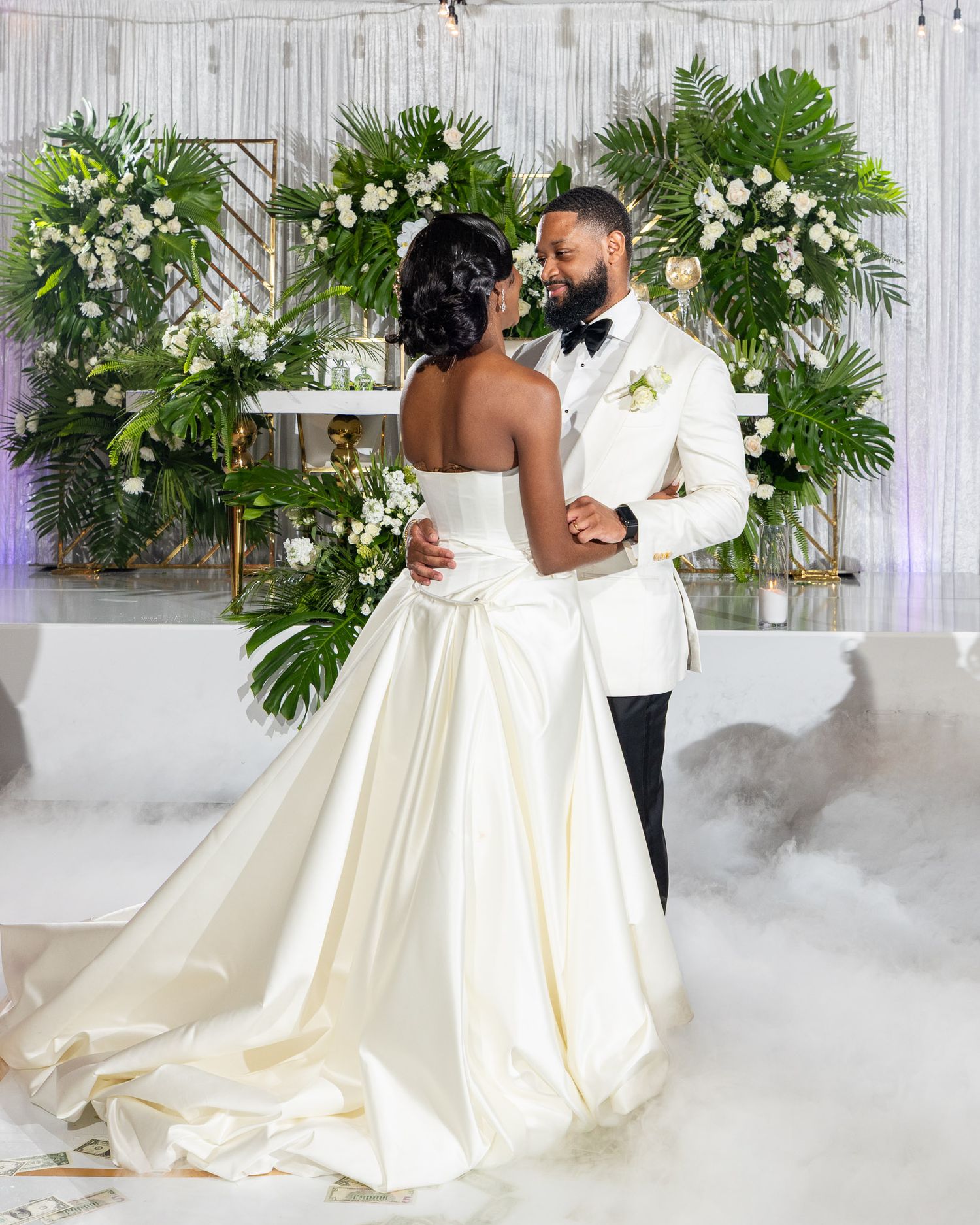 Newlyweds dance surrounded by tropical greenery and white floral arrangements as fog swirls around their feet at reception.