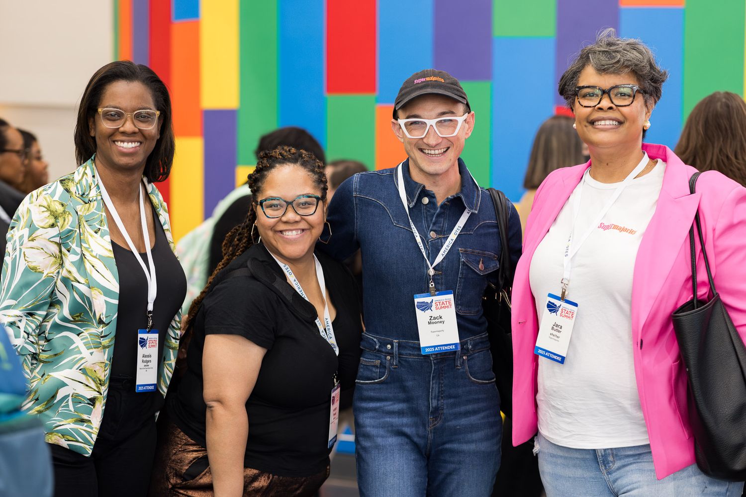 A group of people wearing name tags gather together at a conference with colorful wall decorations in the background.