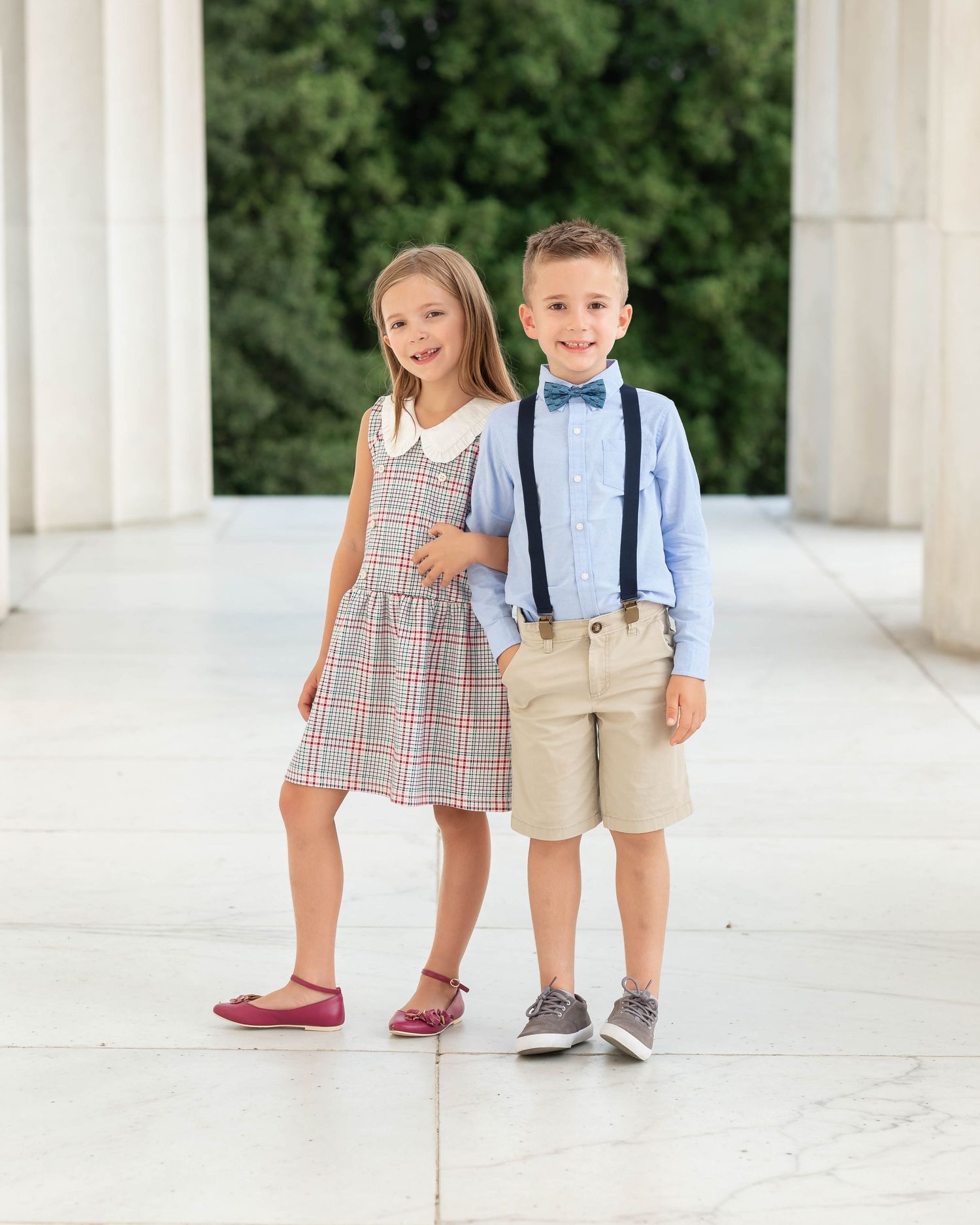 Two young children in dressy outfits pose together, girl in plaid dress and boy in blue shirt with suspenders and bow tie.