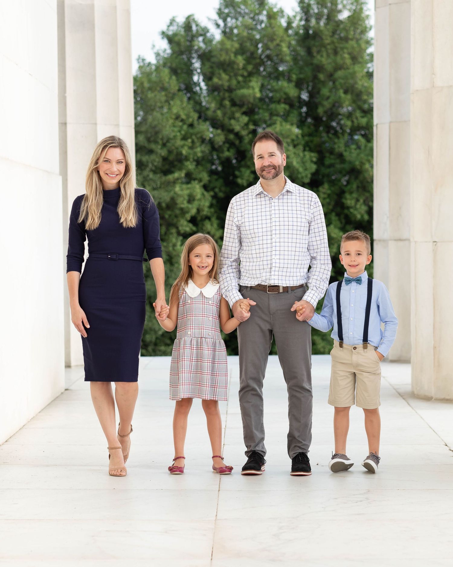 Family of four posing between white columns outdoors, parents holding hands with young daughter and son dressed in formal attire.
