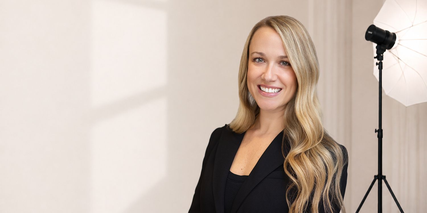 Professional woman with long blonde hair smiling in black blazer, studio lighting setup visible in background.