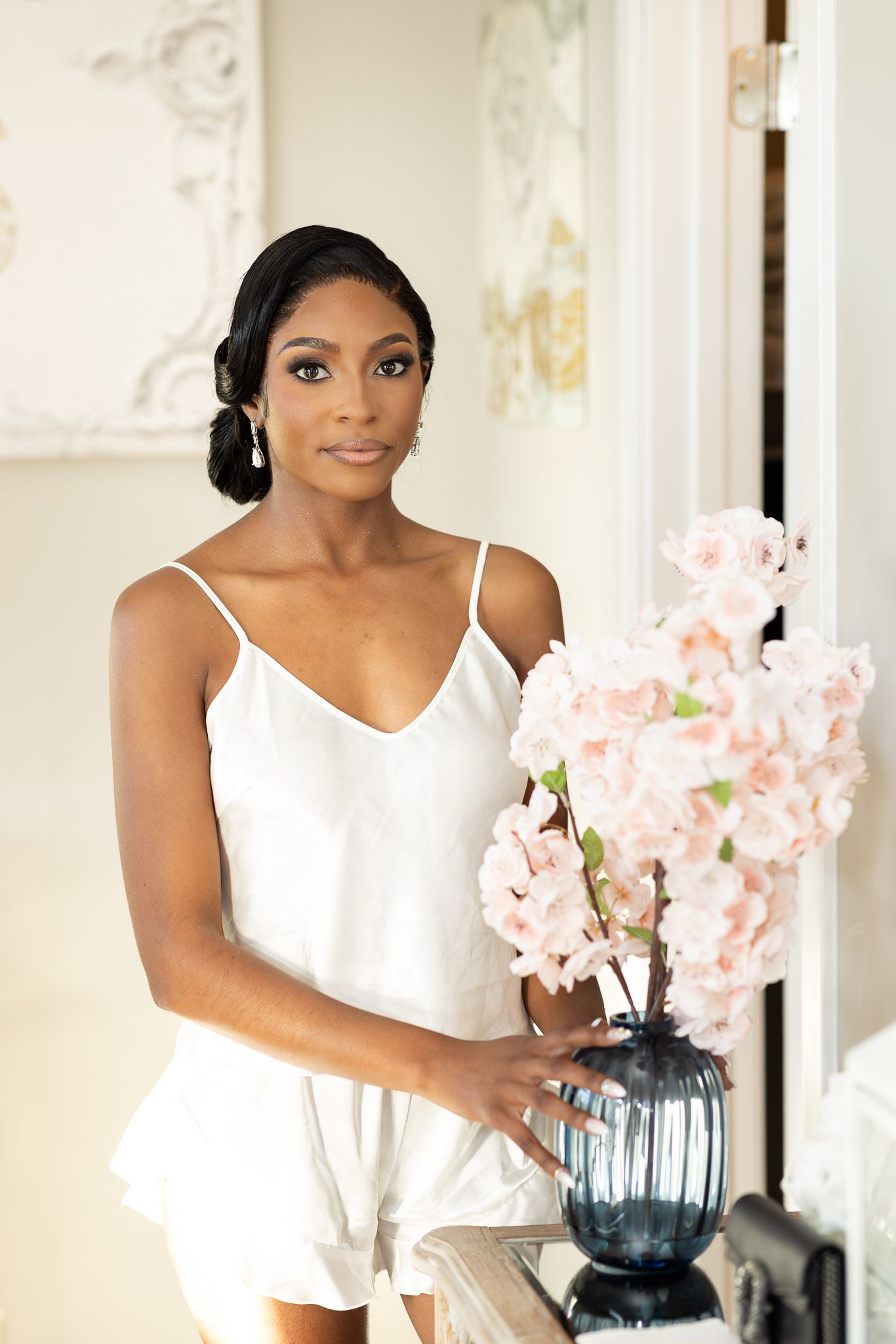 Woman in white dress arranges delicate pink flowers in a blue vase near a bright window.