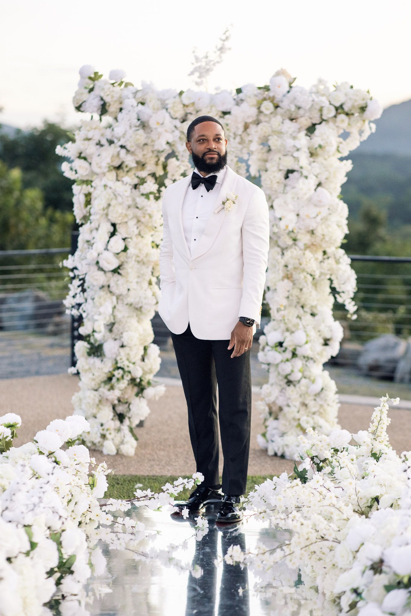 Formal wedding portrait of groom in white dinner jacket near ornate white flower arch.