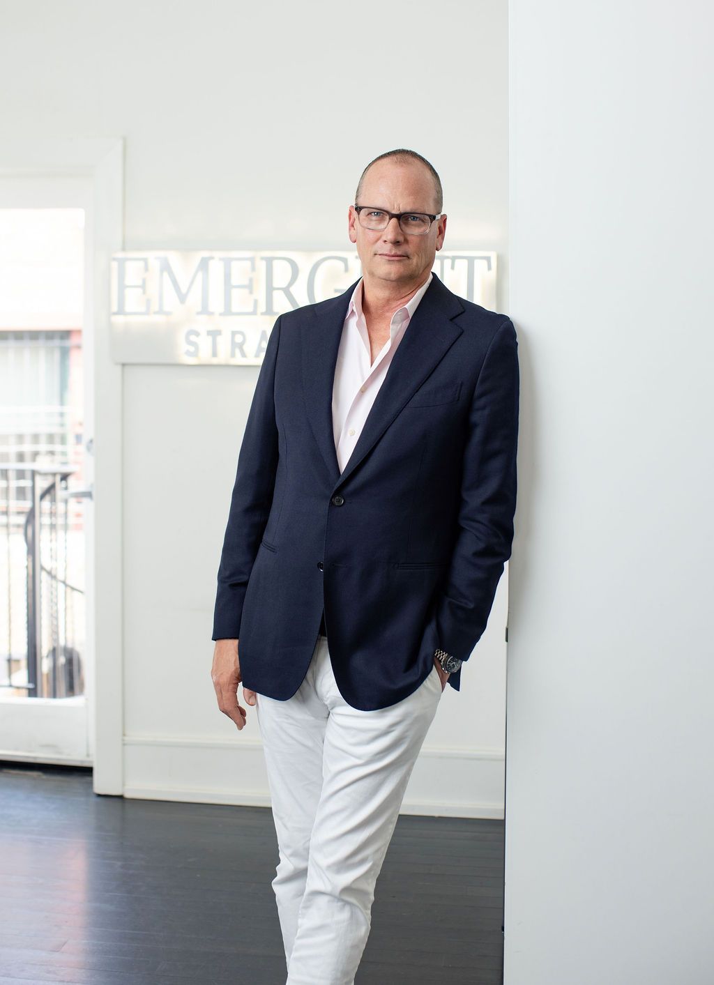 Man in navy blazer and white pants leans against wall at Emergent Strategies office.