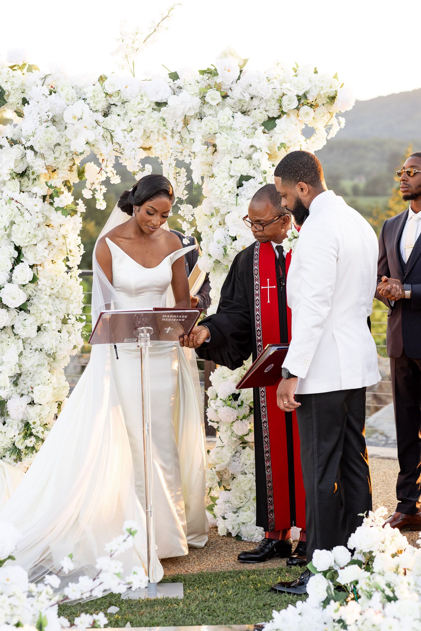 Wedding ceremony under white floral arch with officiant in red stole and wedding party in formal attire.