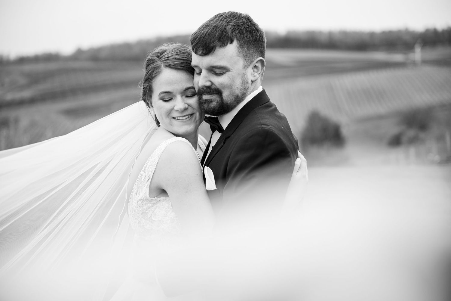 Black and white wedding photo of bride and groom embracing outdoors, veil flowing in the wind with vineyard backdrop.