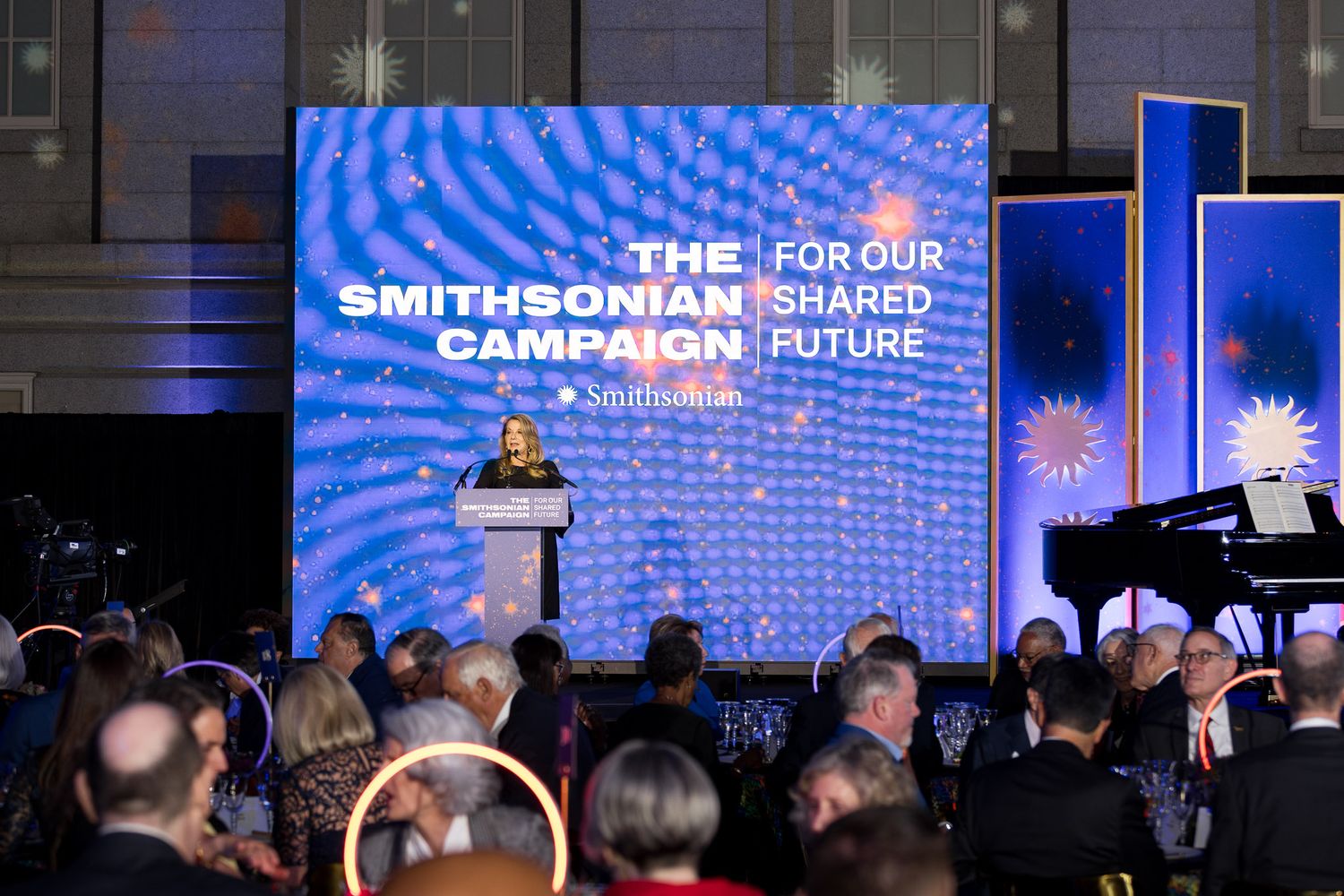 Audience members watch a presentation at the Smithsonian Shared Future event with blue lighting and digital displays.